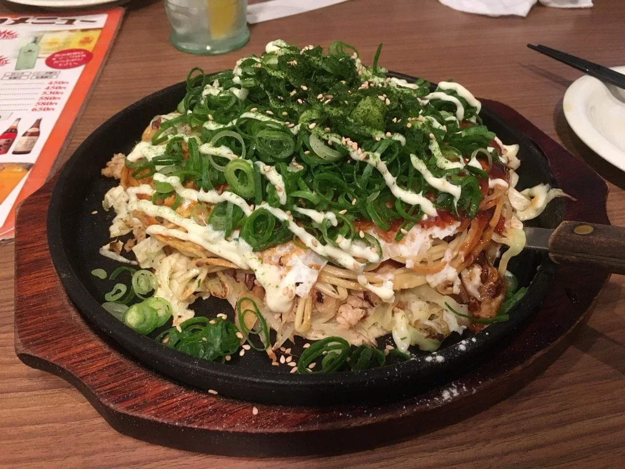 A sizzling plate of chopped vegetables, green onions, sesame seeds, and a creamy white sauce on top, served on a wooden tray.