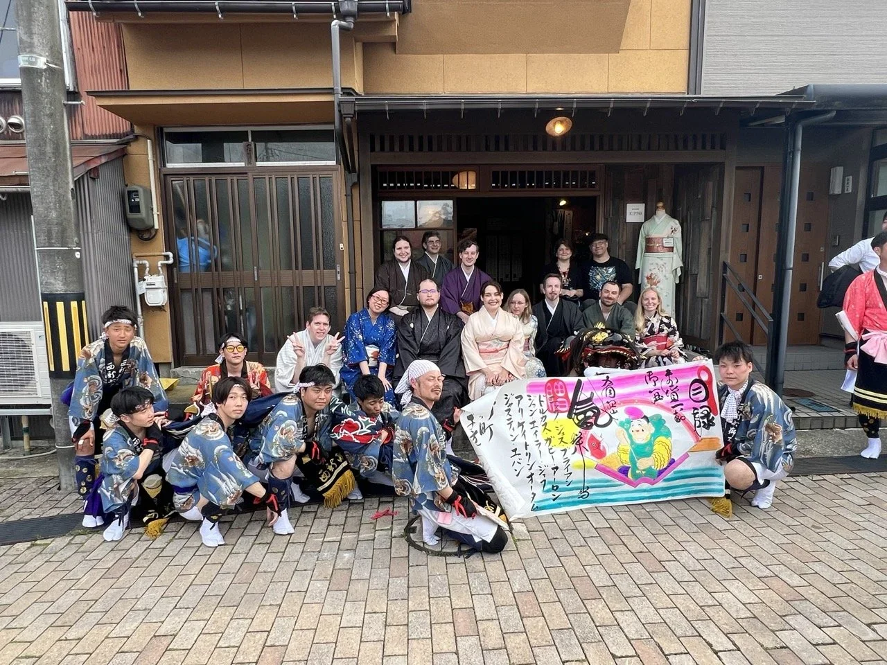 Group of people wearing traditional Japanese festival costumes, some holding a colorful banner with Japanese writing, posing outside a building with a wooden facade. A mannequin dressed in kimono can be seen in the background.