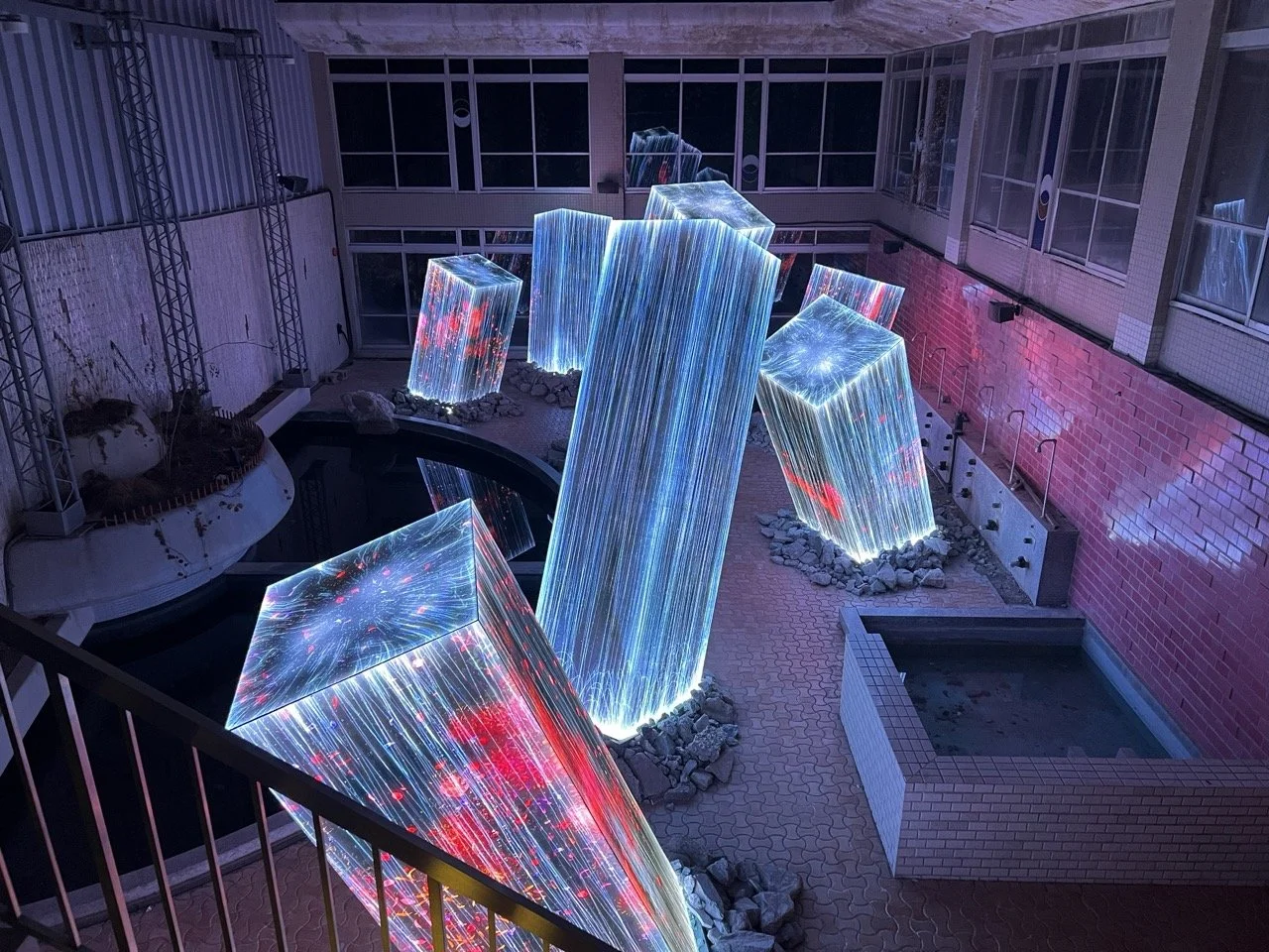 Massive digital screens cover giant obelisks rising from the broken tiles and scenery of an abandoned bath house.