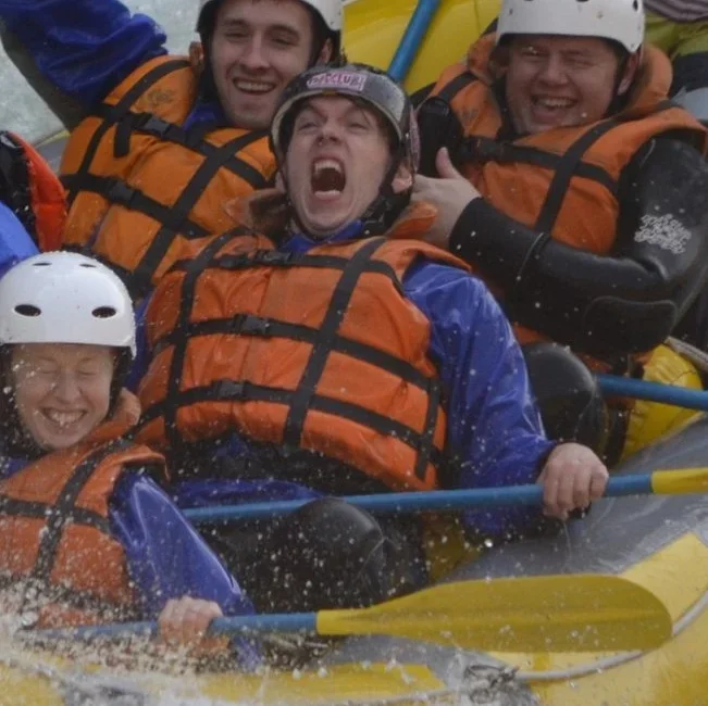 Group of people white water rafting with life jackets and helmets, smiling and enjoying the river splashes.