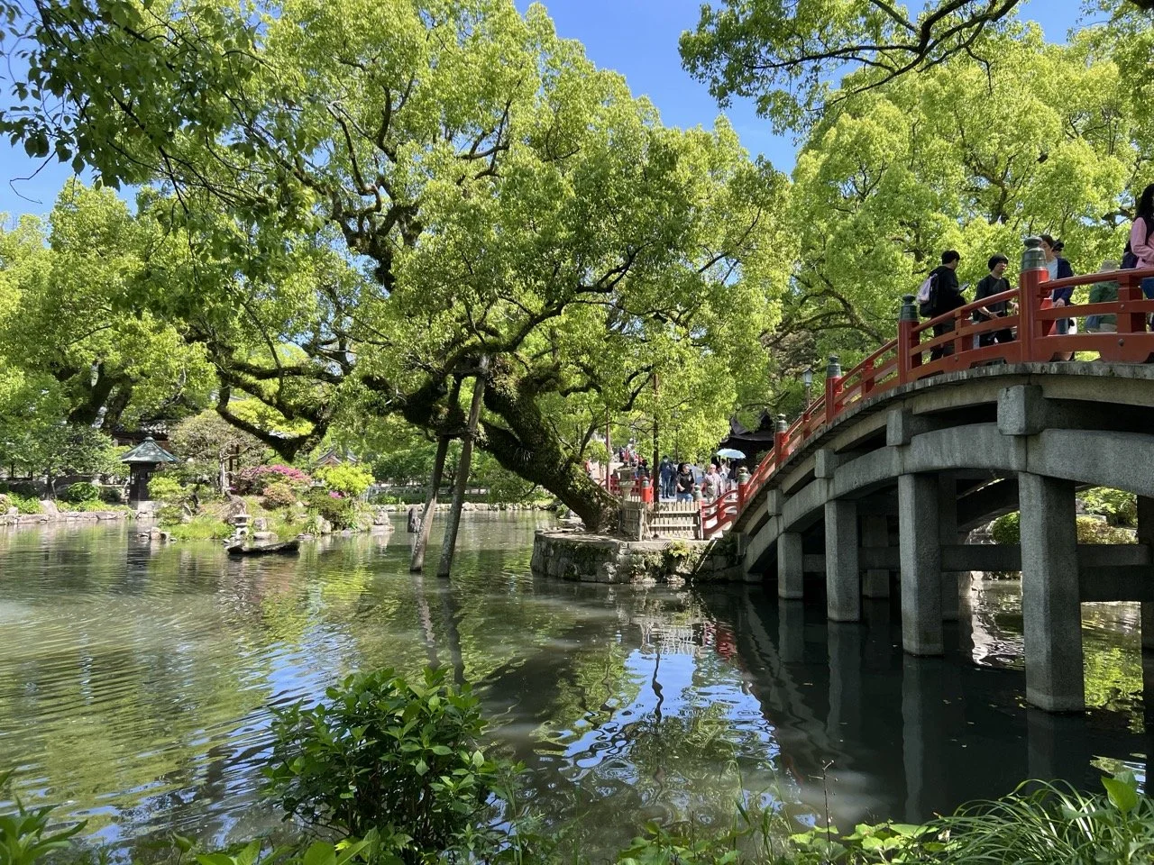A bright red arched bridge with people traveling across it leads people through a collection of tree-lined islands over a collection of ponds.