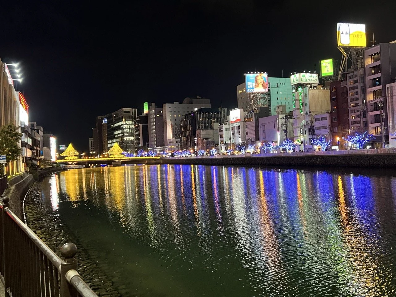 A massive river runs through a huge city at night, as surrounding buildings cast neon light reflections off the water.