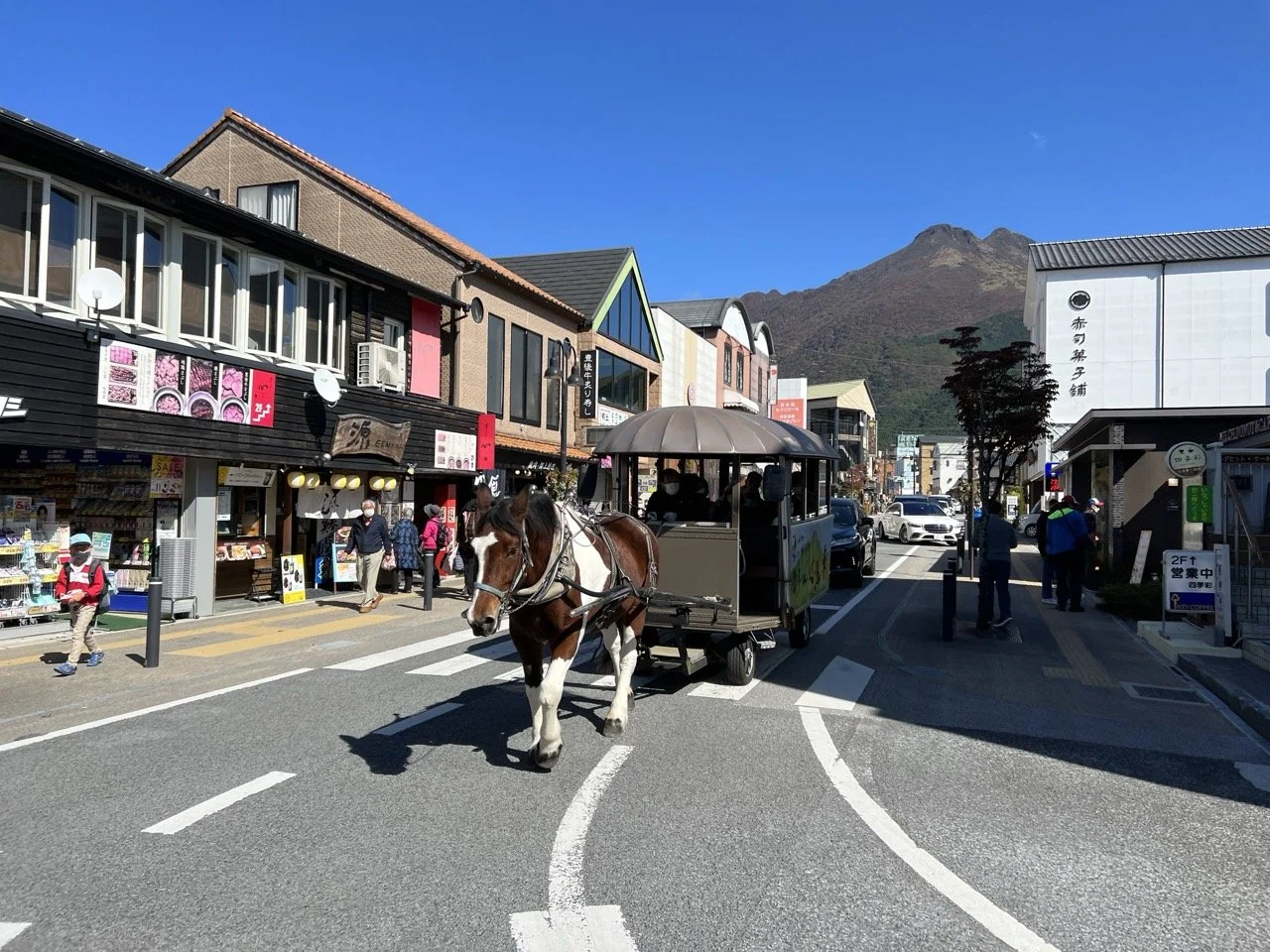 A horse pulls a cart full of people down a scenic street lined with shops. A mountain towers over the scene in the background.