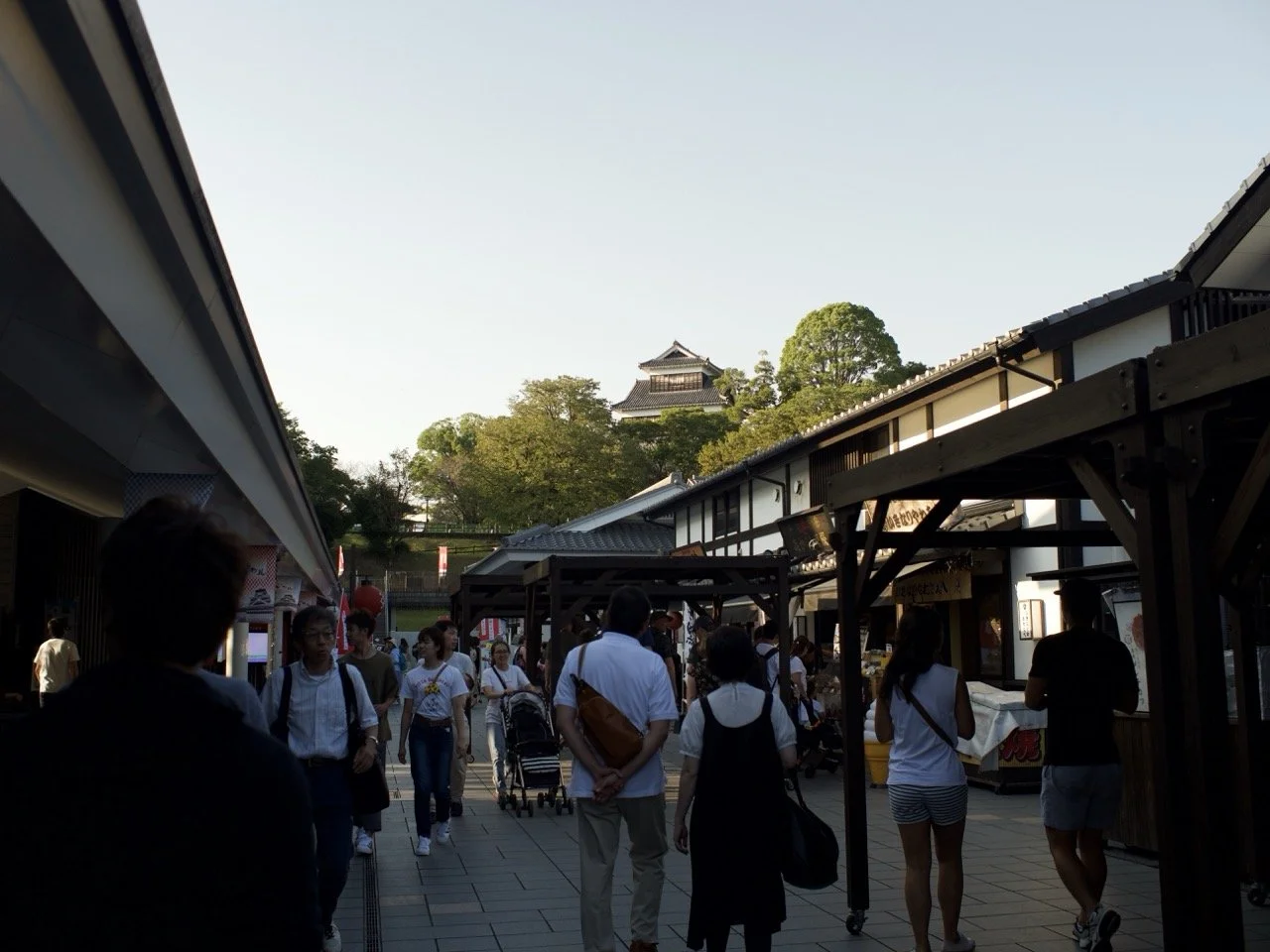 An outdoor shopping plaza bustling with people in the afternoon sunshine. The top of Kumamoto Castle can be seen in the background.