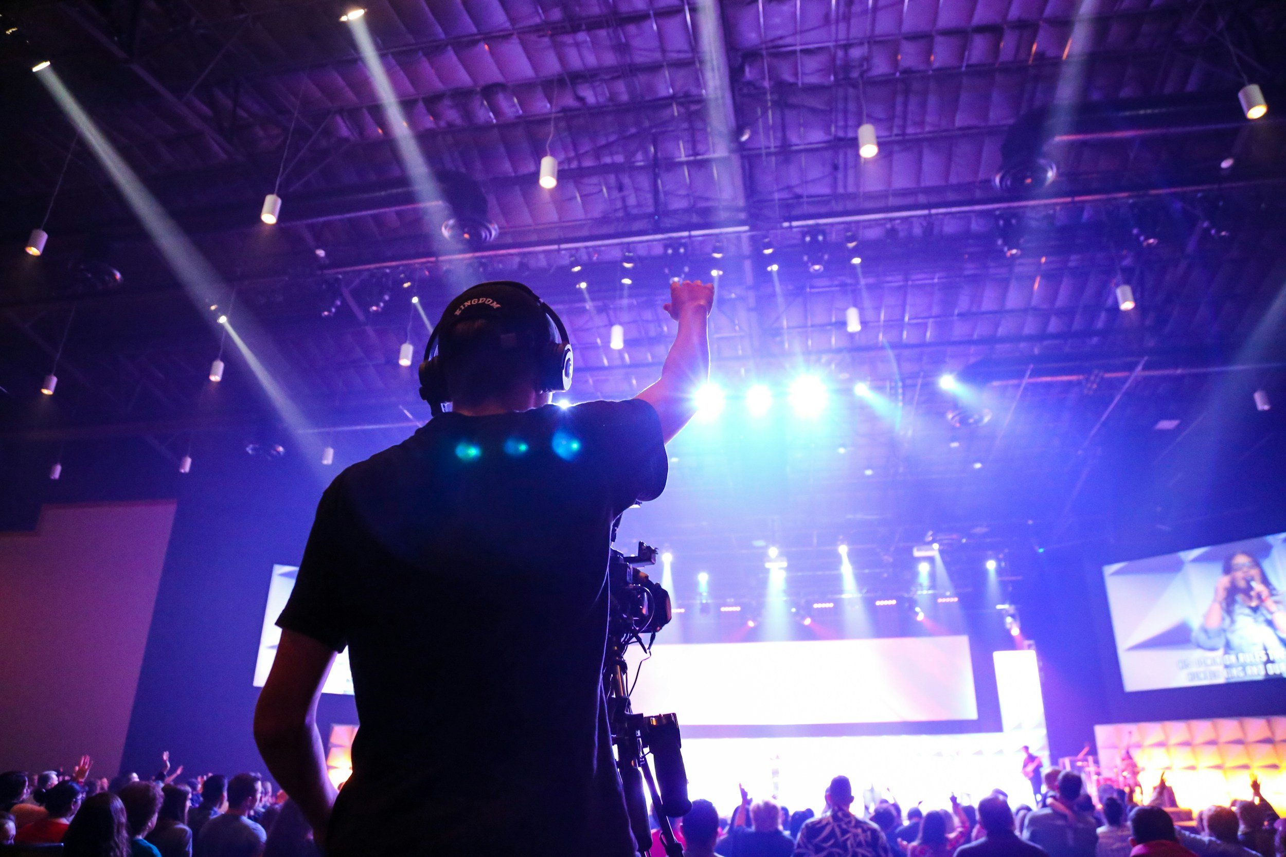 A person with a headset and camera at a concert or conference, raising a fist towards bright stage lights in a large indoor venue with an audience.
