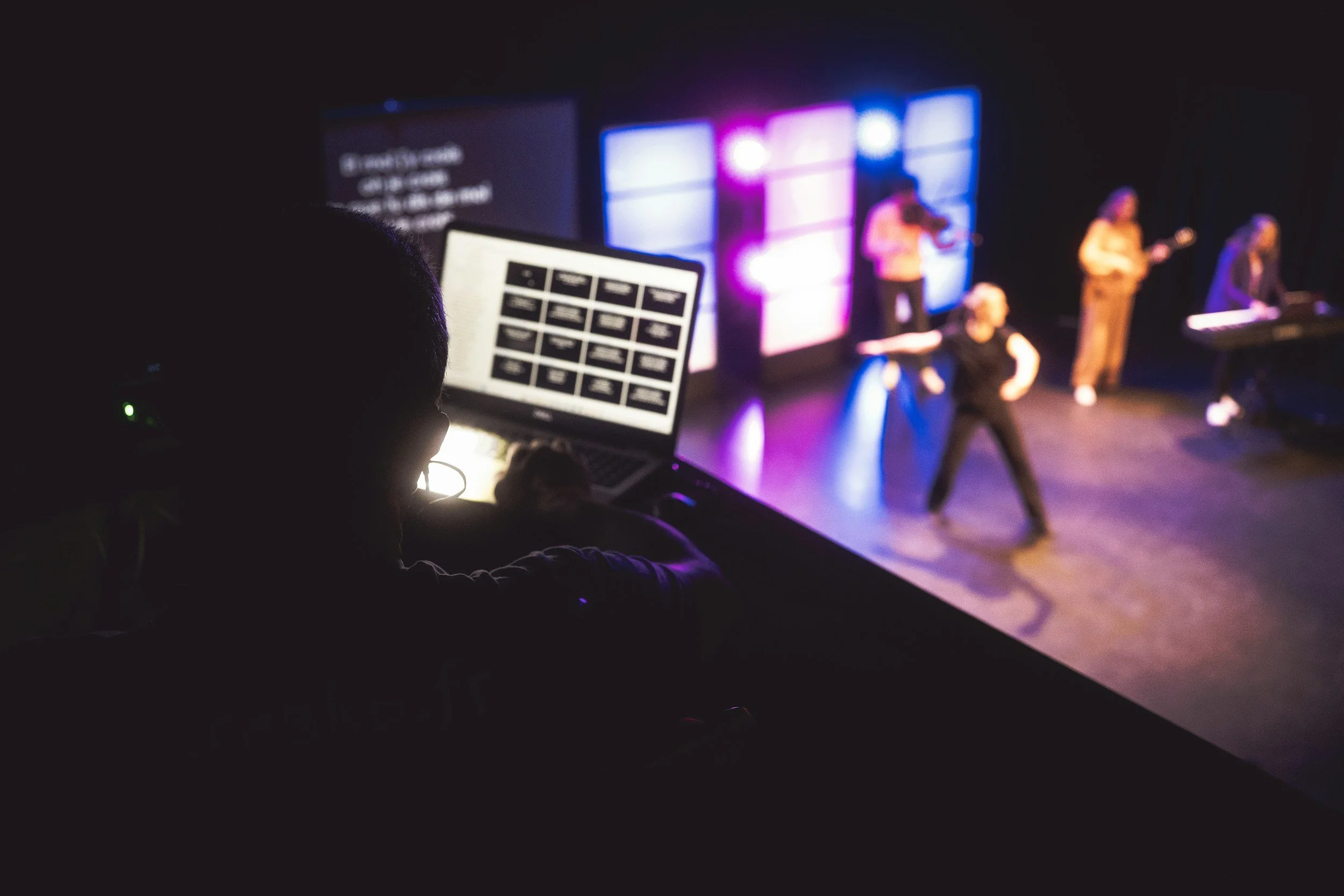 A control booth with a person working on a laptop, observing performers on stage with colorful lighting and video screens in the background.