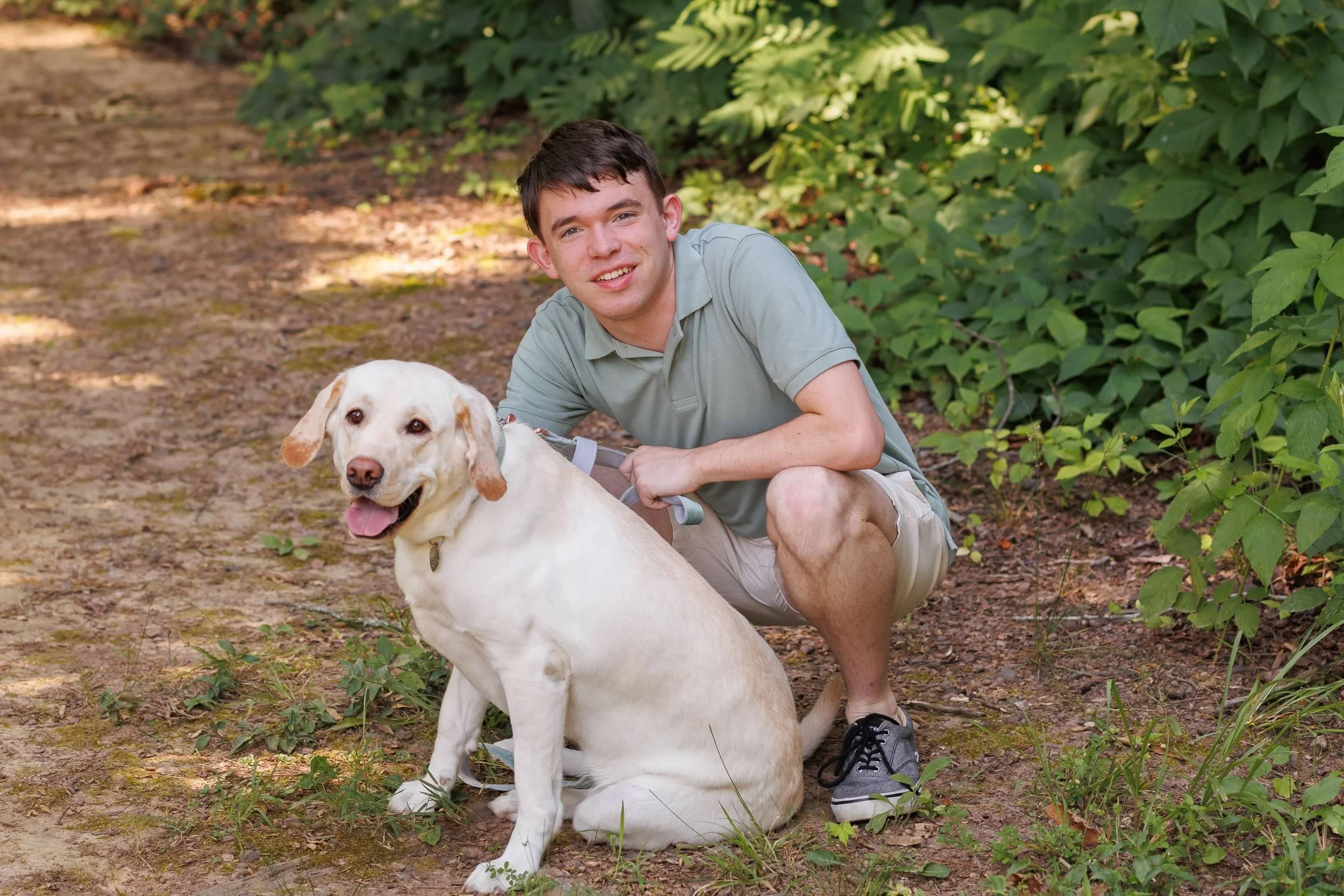 A young man with short dark hair, wearing a gray polo shirt and beige shorts, kneeling on a dirt path in a wooded area, holding a large white Labrador Retriever dog with floppy ears and a happy expression.