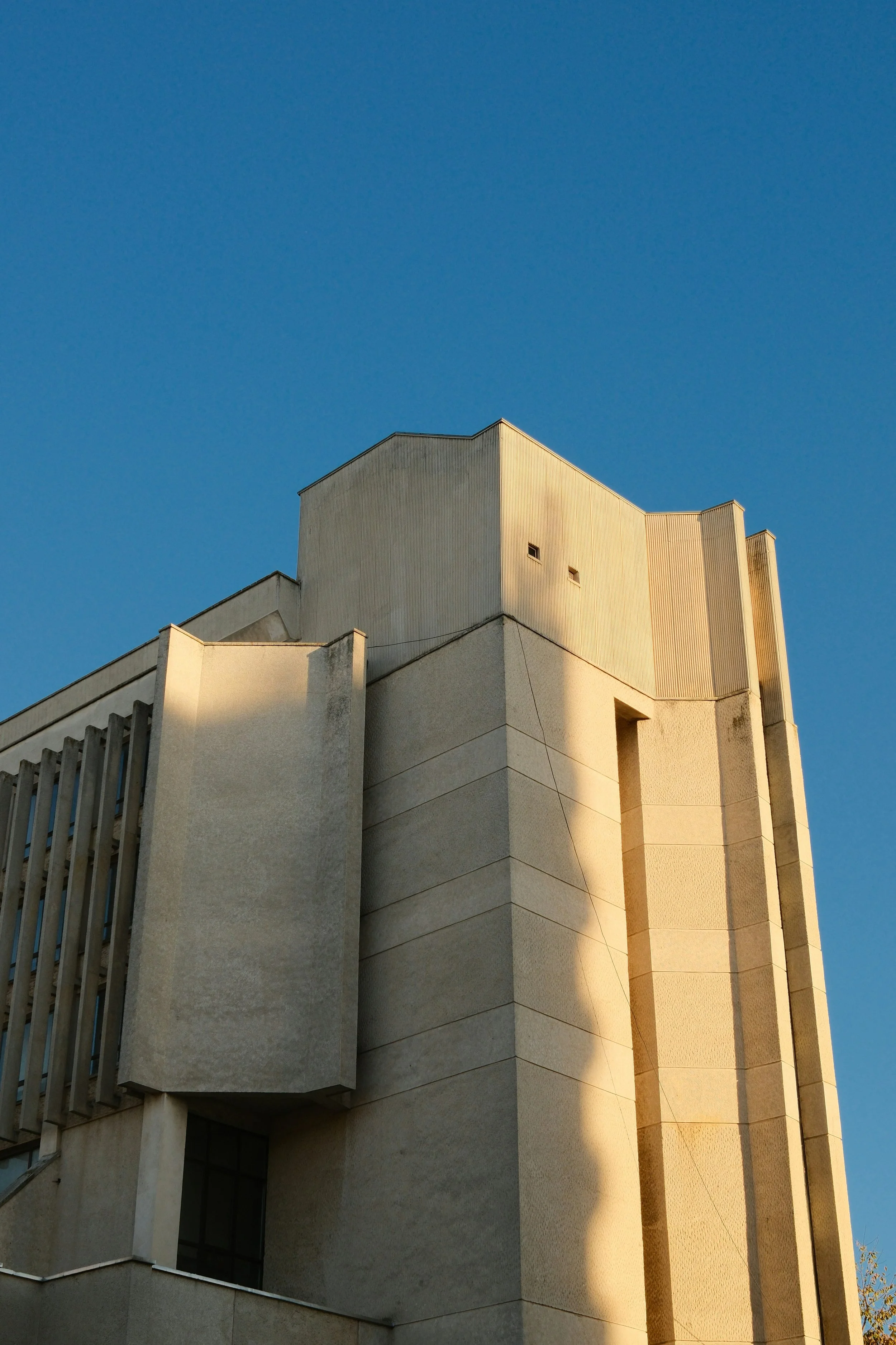 A tall, modern beige building with sharp angles and vertical lines, illuminated by sunlight, under a clear blue sky.