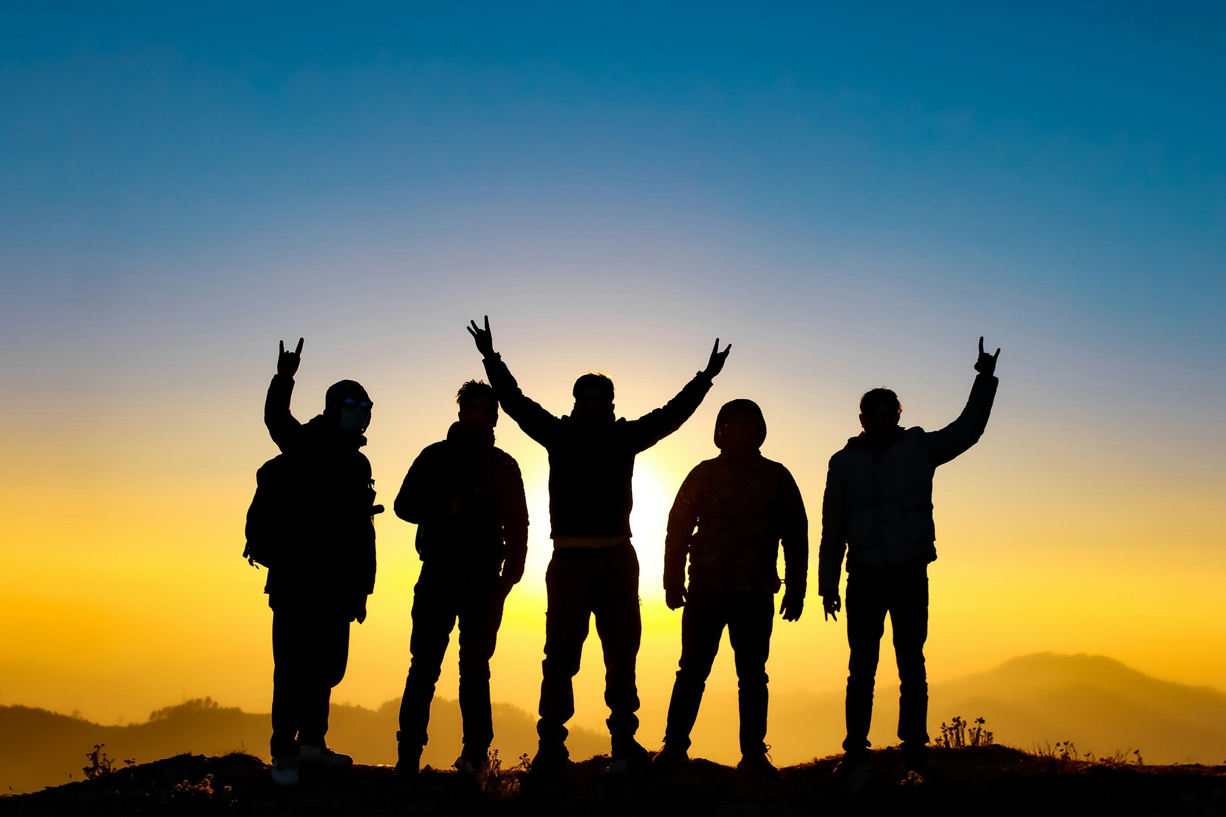Silhouetted group of five people standing on a hill at sunset, some with arms raised and making peace signs.