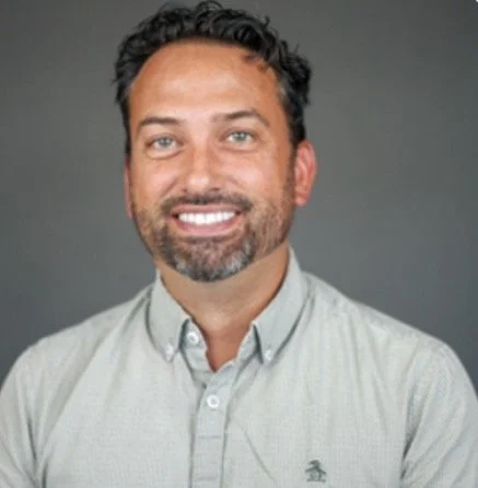 Headshot of a smiling man with dark hair and beard, wearing a light-colored button-up shirt, against a gray background.