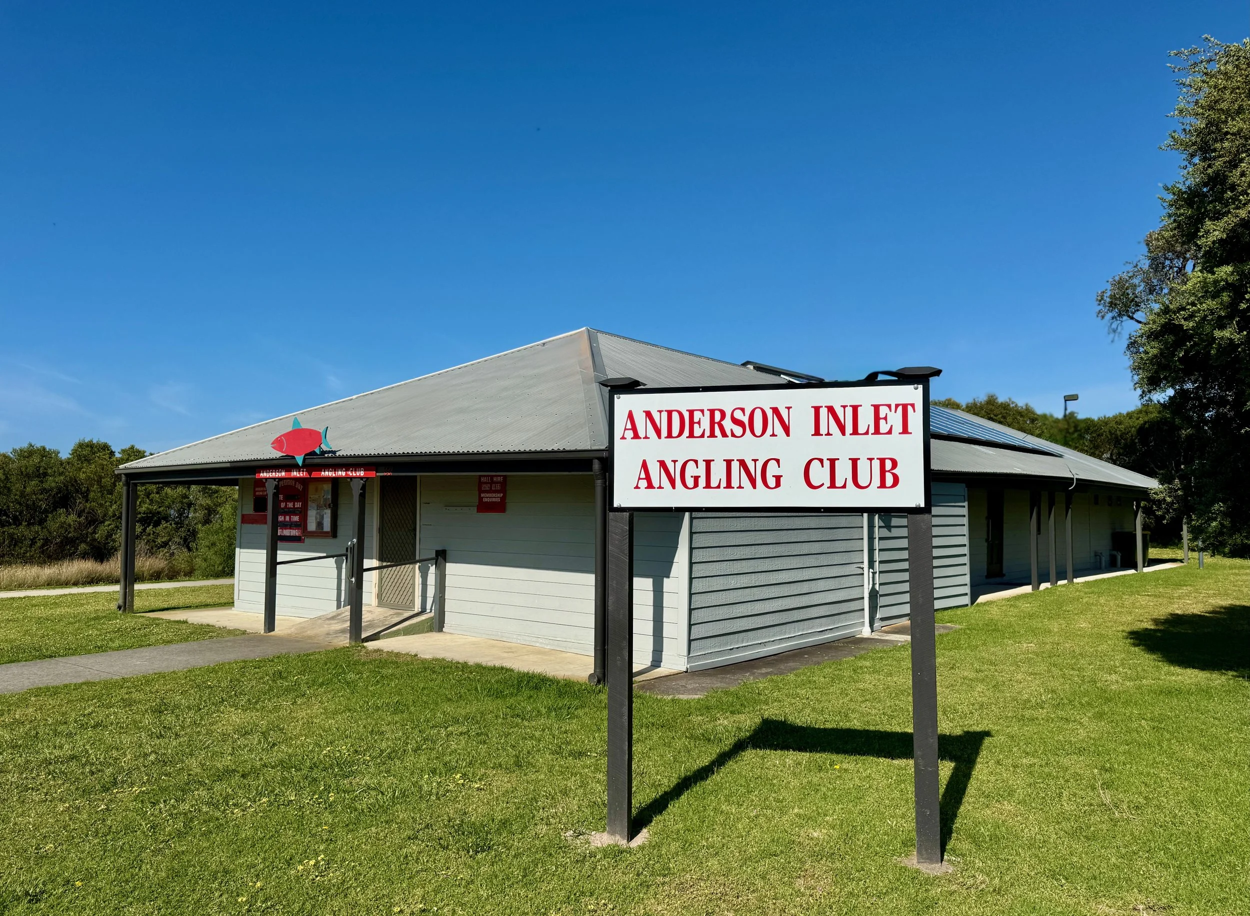 A building with a sign that reads 'Anderson Inlet Angling Club' and a logo of a fish mounted on the front. The building has grey siding, a metal roof, and is surrounded by green grass and trees under a clear blue sky.