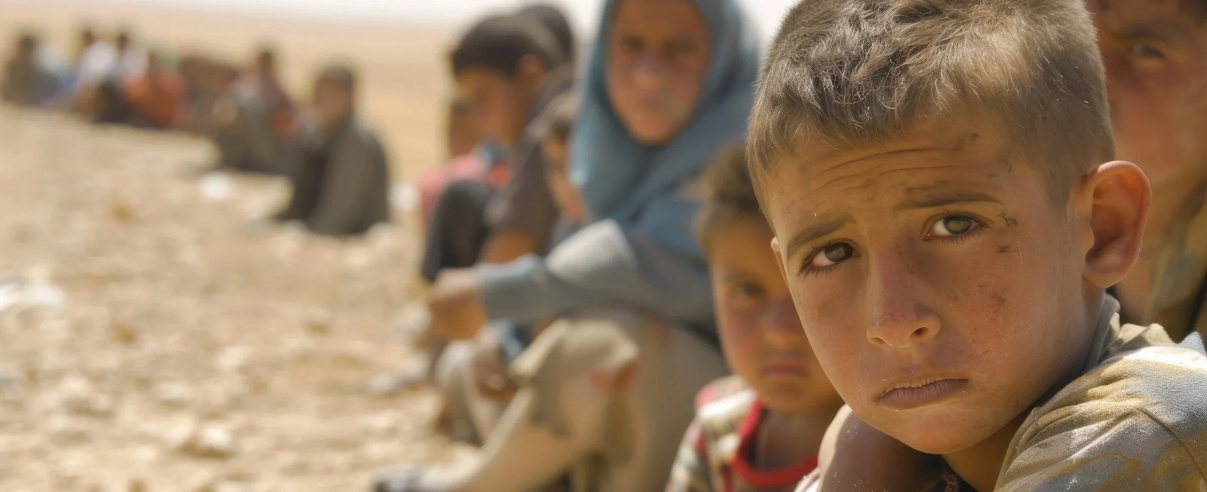 A line of children sitting on the ground, with the focus on a young boy in a dusty outdoor setting.