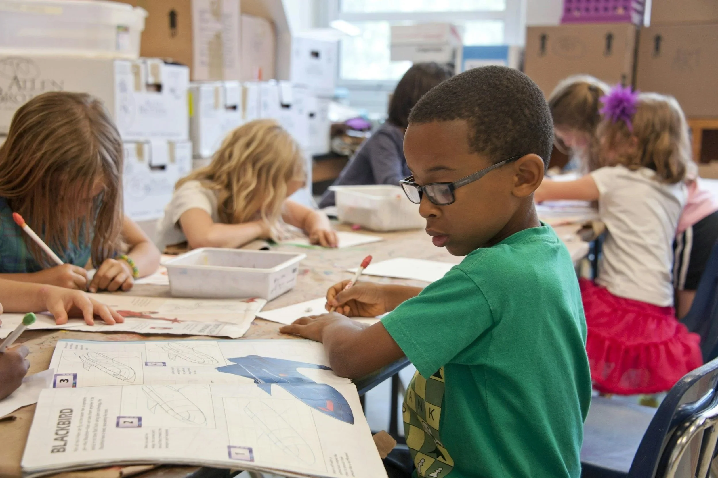 A group of children sit at a table in a classroom, working on coloring and drawing activities with worksheets and colored markers.