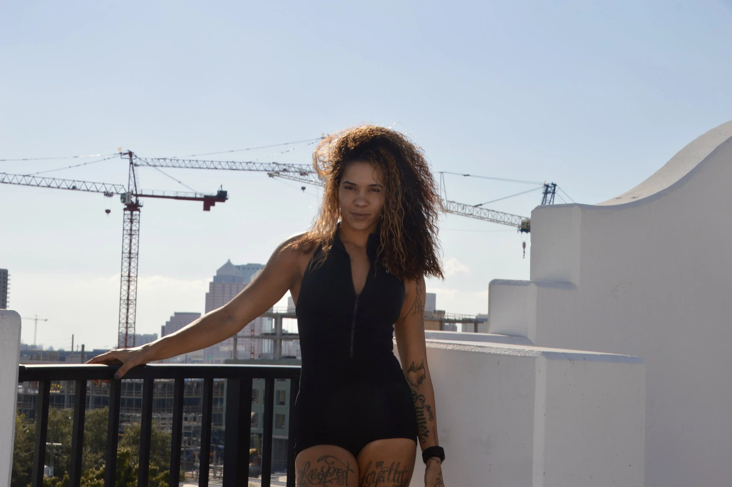 Woman with curly hair standing on a rooftop terrace with city buildings and cranes in the background.