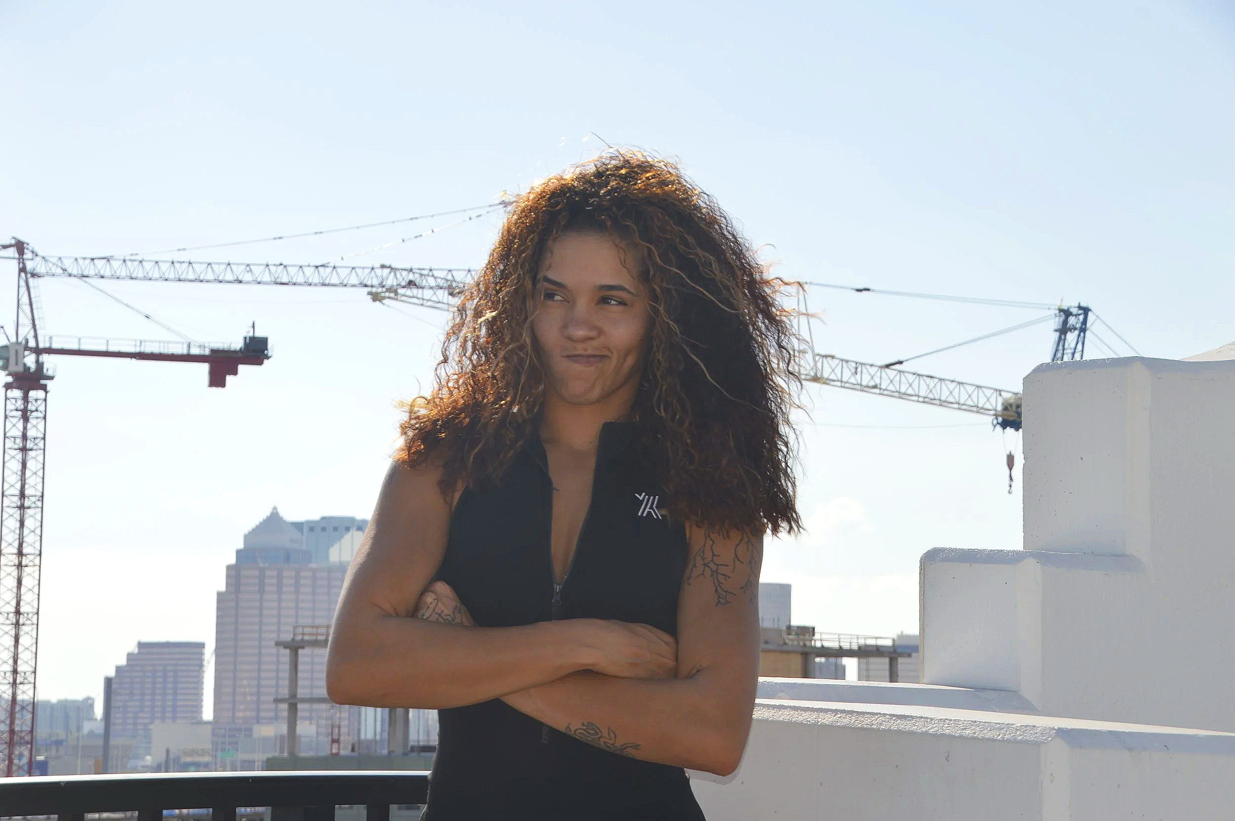 A woman with curly hair and tattoos on her arms stands with arms crossed on a rooftop with a city skyline and construction cranes in the background.
