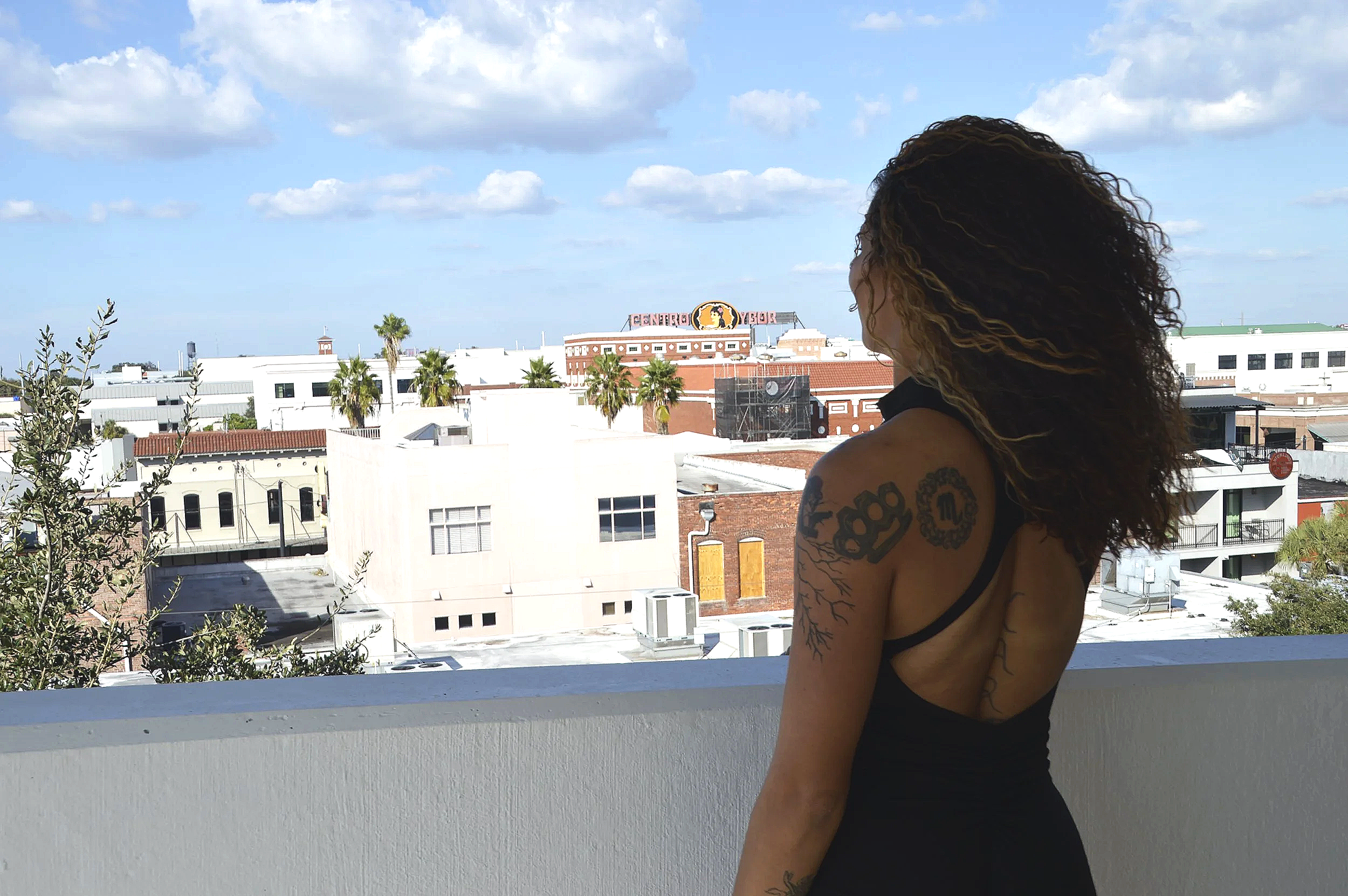 A woman with tattoos on her arm and back, wearing a black sleeveless top, standing on a balcony overlooking a cityscape with palm trees and buildings under a partly cloudy sky.