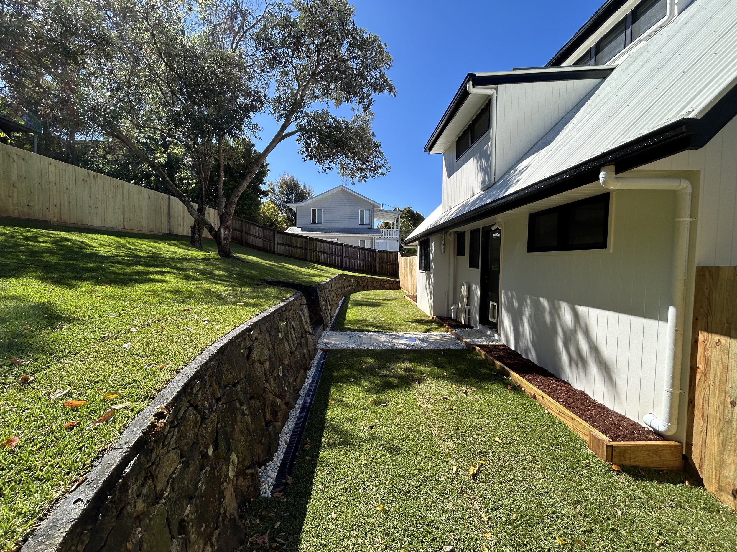 Side yard of a house with a stone retaining wall, a grassy slope with a tree, and a white house in the background under a clear blue sky.