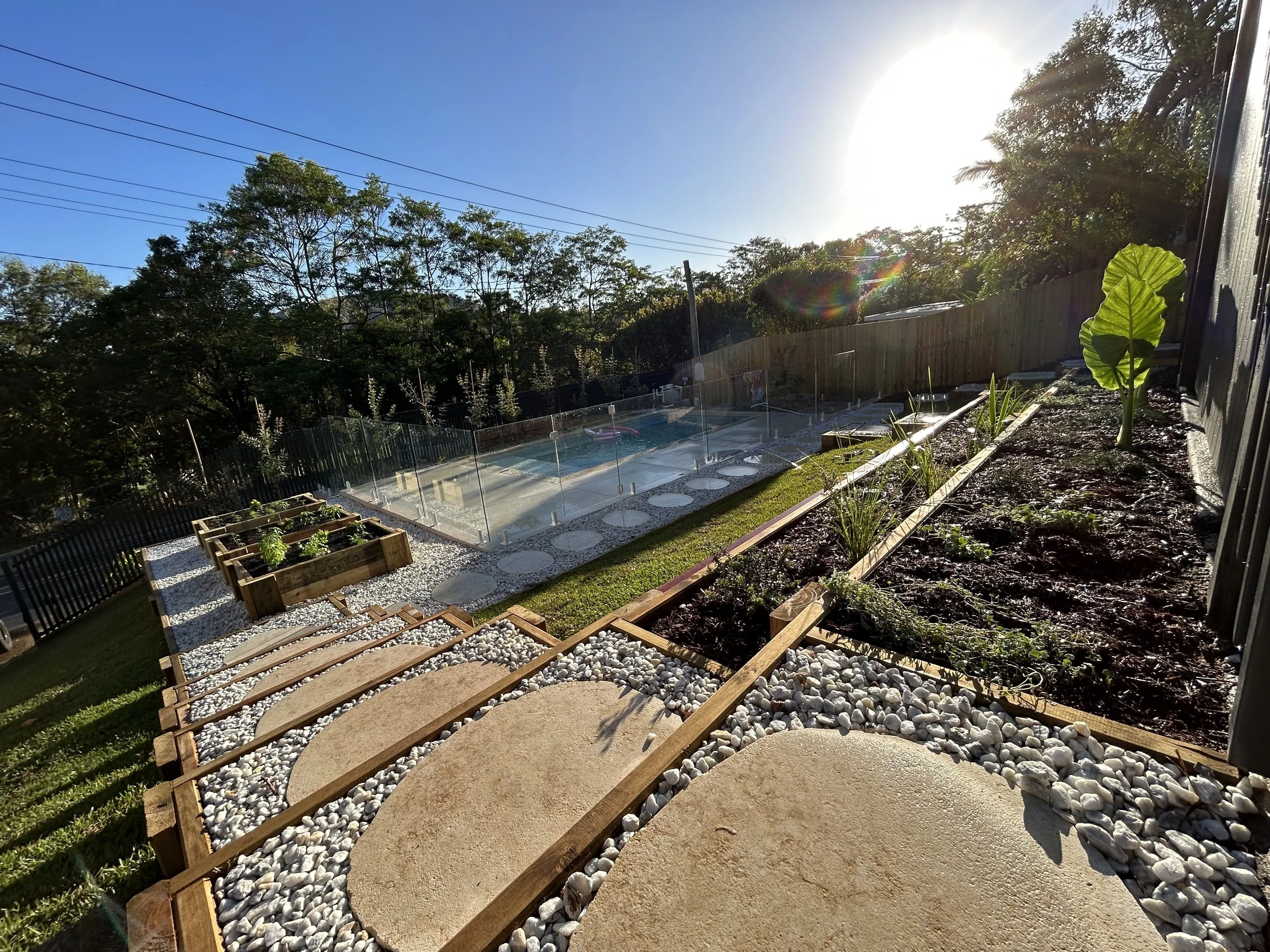 Backyard garden with vegetable plots, a pool enclosed by glass fencing, stone pathway, and various plants under a sunny sky.