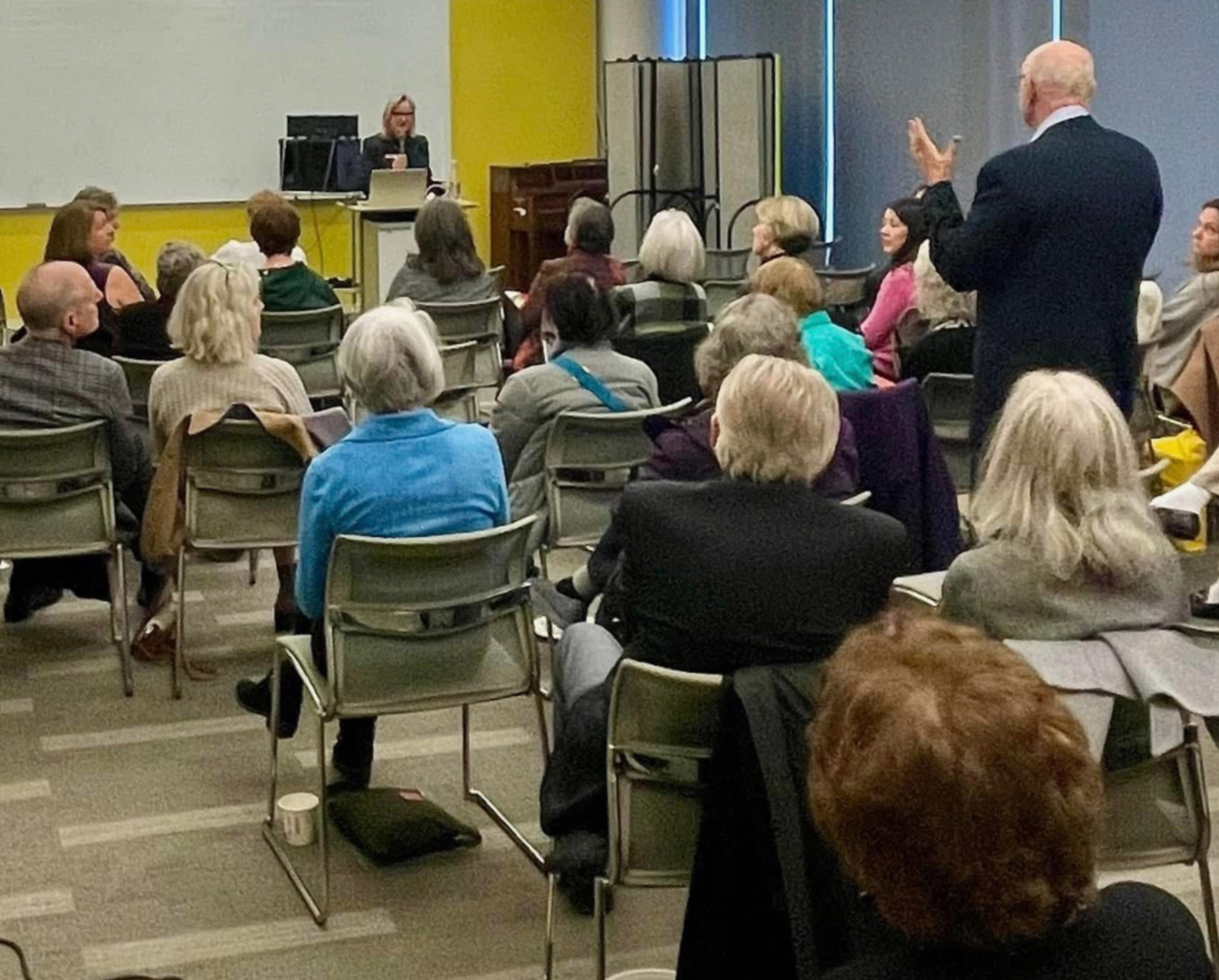 An indoor conference room filled with seated people listening to a speaker with a microphone. A man stands and raises his hand, possibly asking a question. The room has a yellow and blue wall, with a woman at a podium in front of the audience.