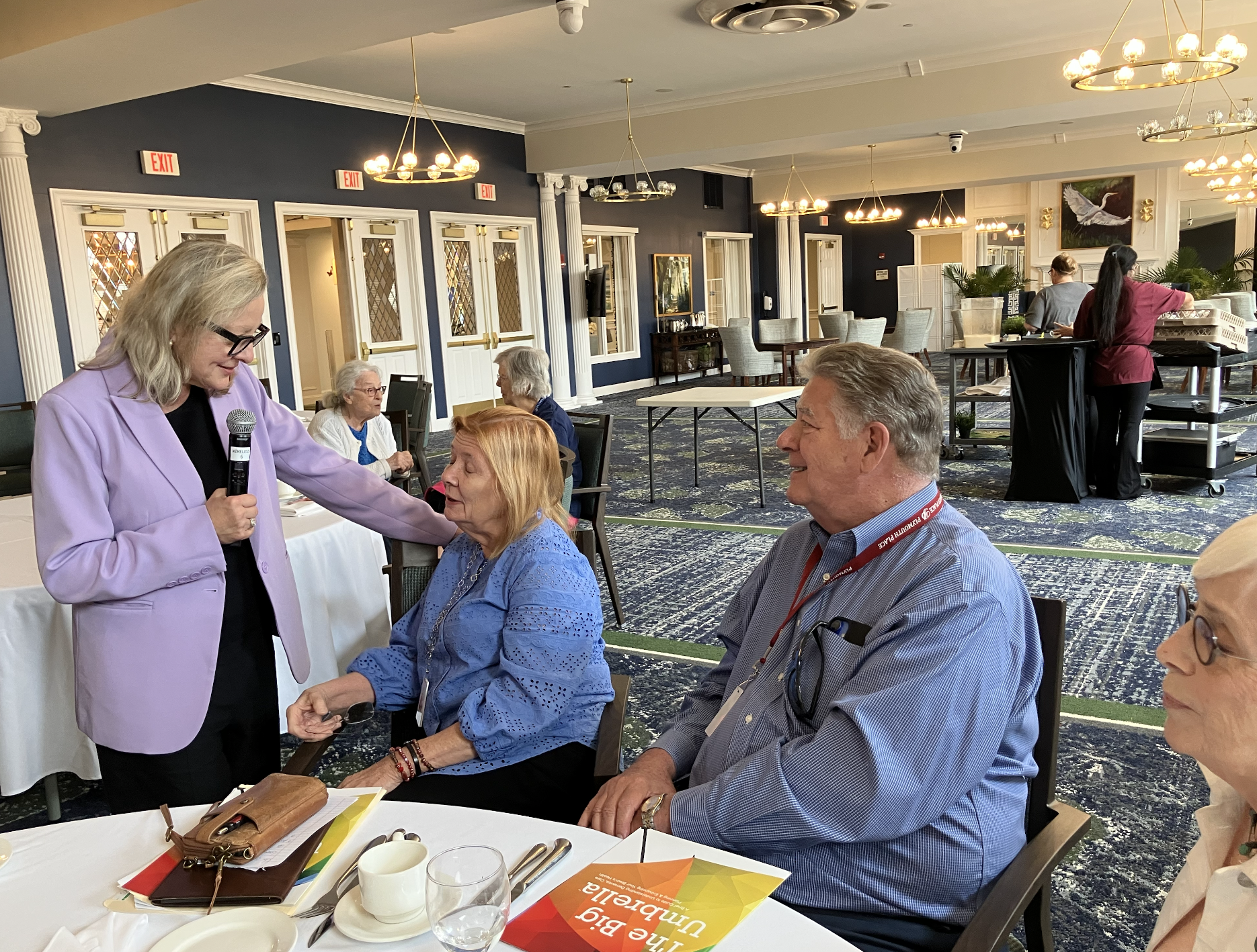 A woman in a lavender blazer holding a microphone and talking to an elderly woman in a blue blouse during a social gathering in a decorated dining room.