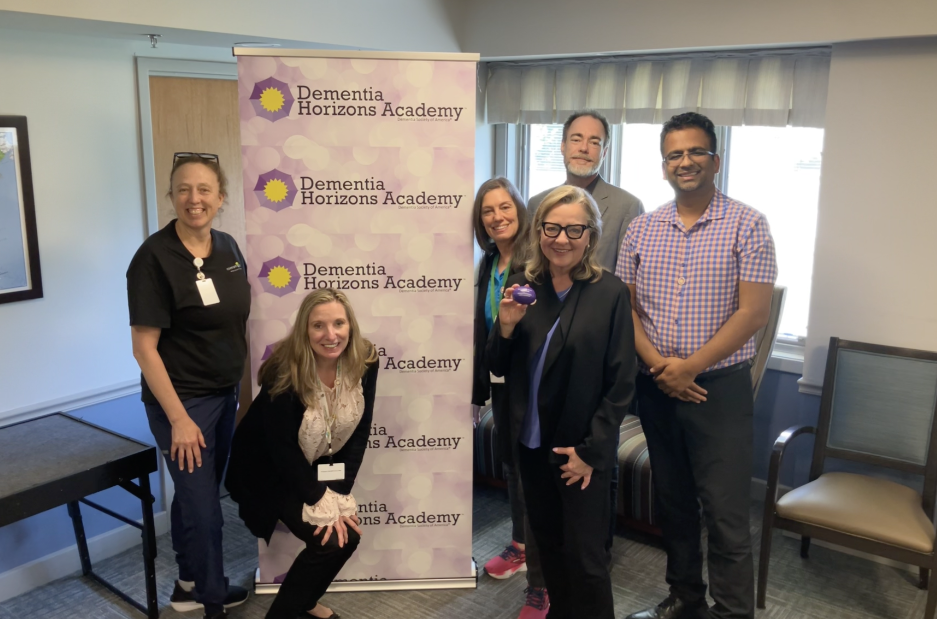 Group of six people smiling and posing for a photo in front of a Dementia Horizons Academy banner during an event or meeting, indoors with window light.