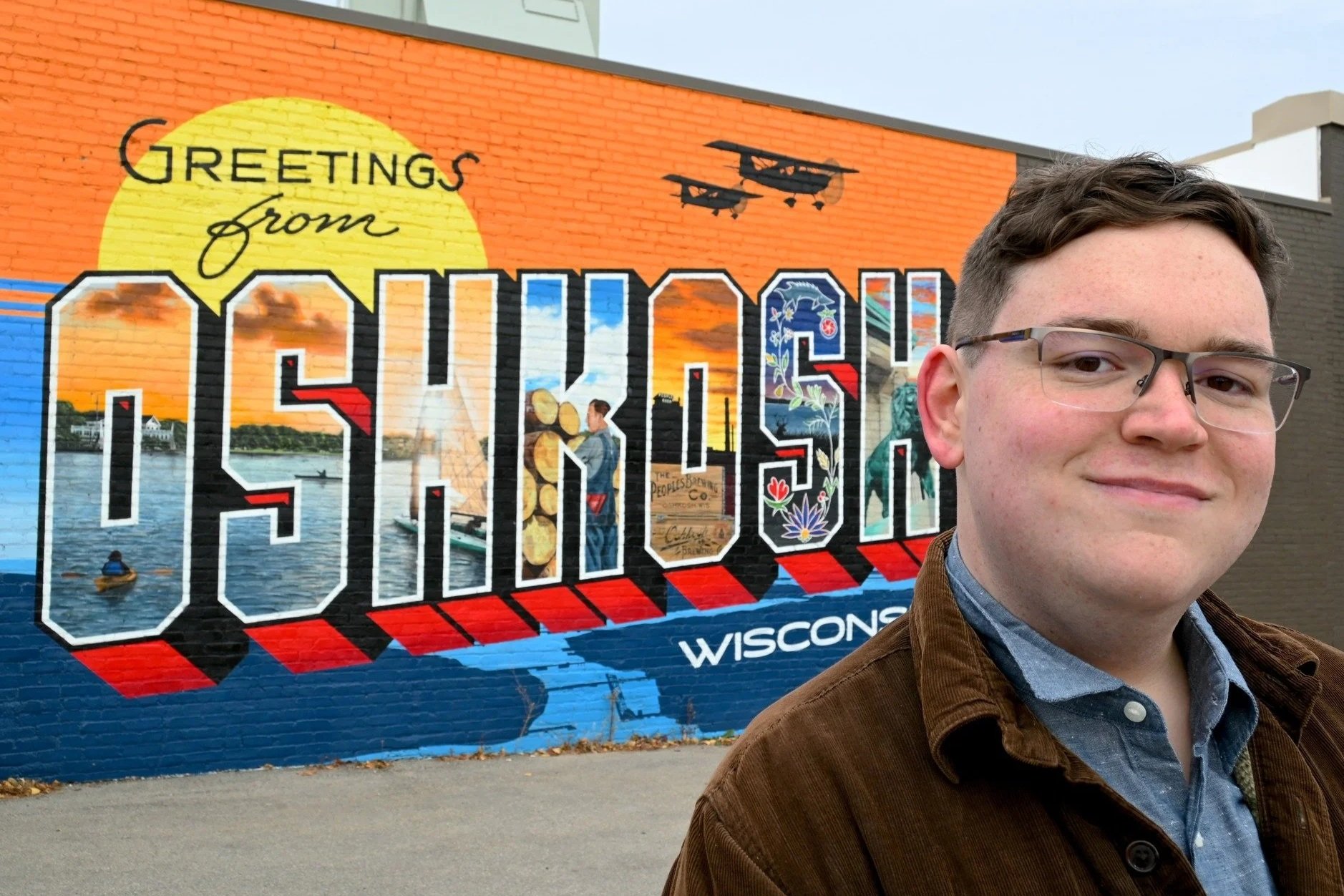 A young man with glasses and a brown jacket stands in front of a colorful mural that reads 'Greetings from Wisconsin' with a lake scene, boats, and planes painted on a brick wall.