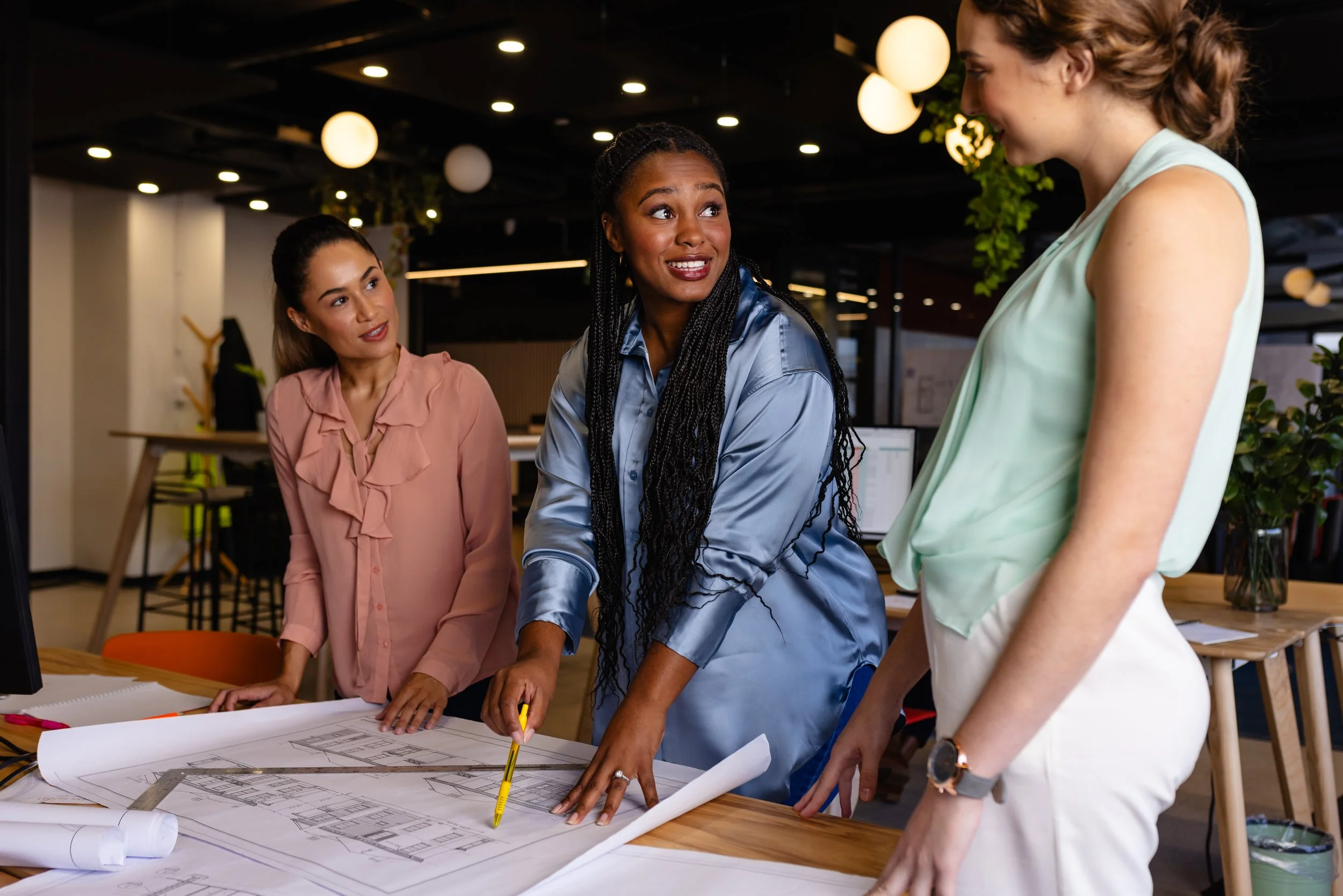 Three women collaborating on a construction or architectural project at a table with blueprints in an office setting.