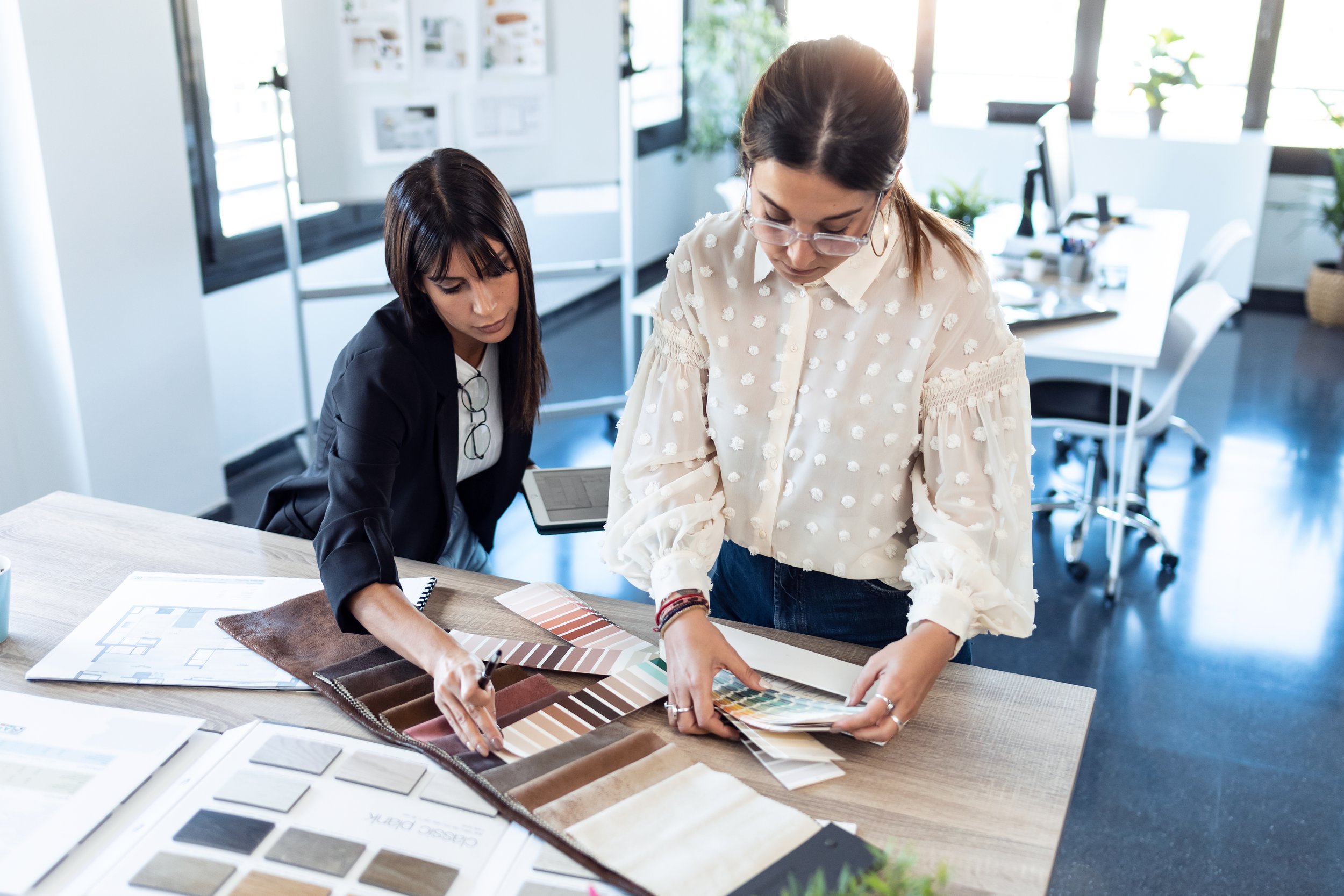 Two women working on interior design, examining fabric and color samples at a modern office table.