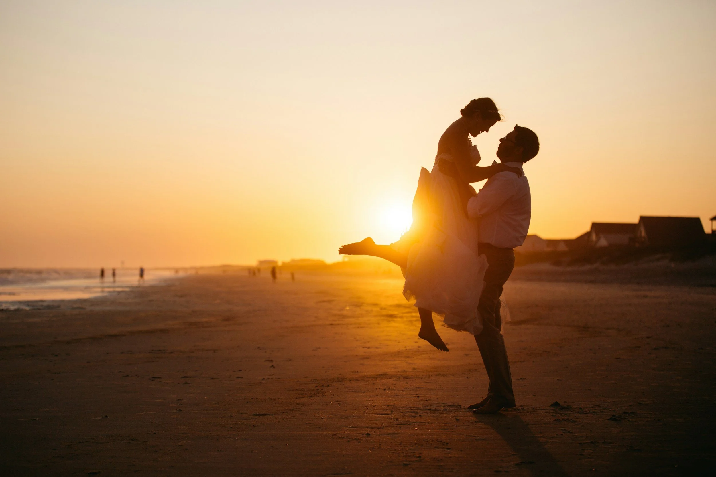 A couple on a beach at sunset, with the man lifting the woman who is sitting on his arms. The scene is silhouetted against the orange sky, with gentle waves and distant houses visible in the background.