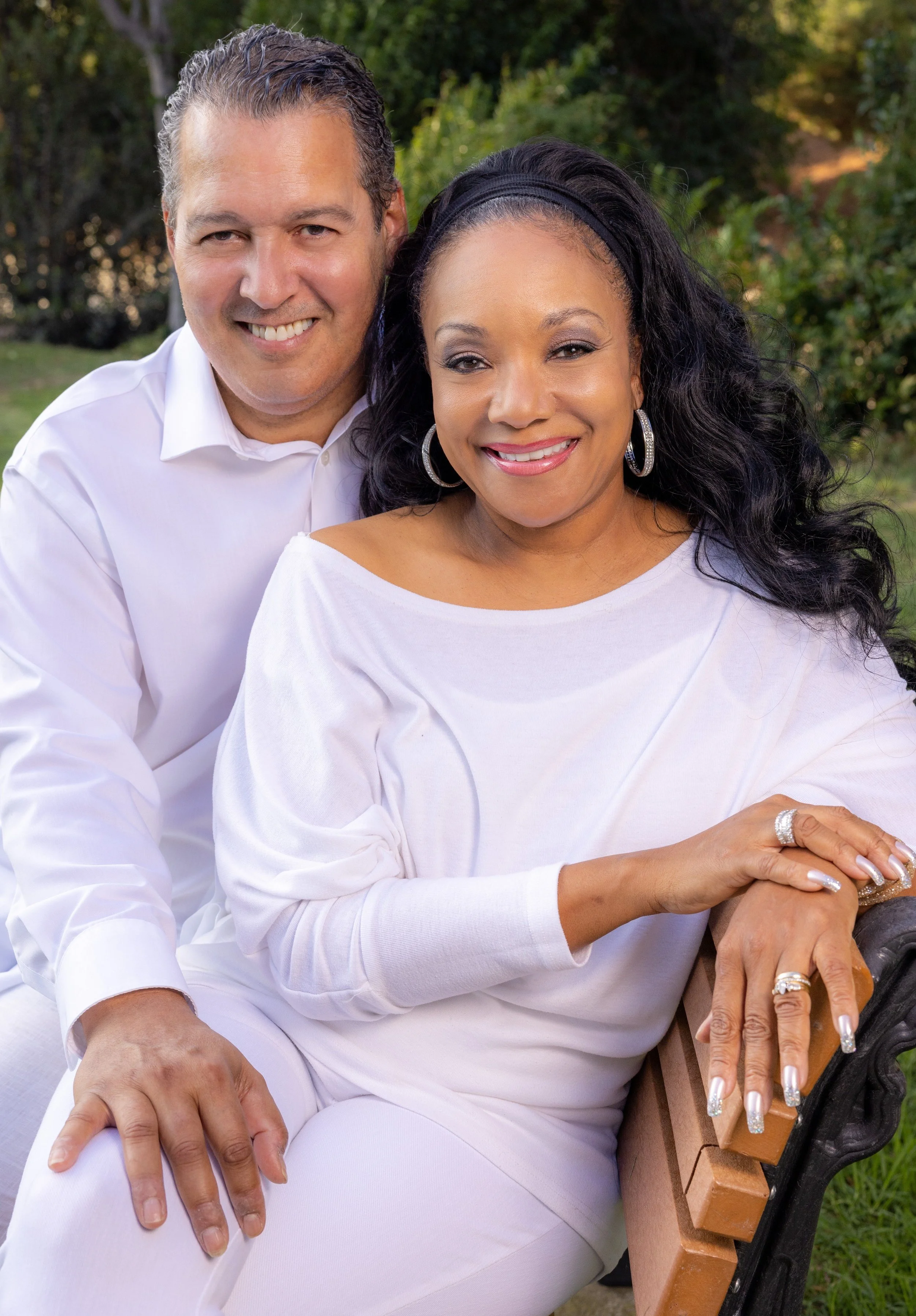 A smiling couple sitting on a park bench outdoors, with a green, leafy background. The woman is wearing a white top, silver hoop earrings, and rings, and the man is in a white shirt. They are close together, with the woman resting her hands on the bench, and both are looking at the camera.