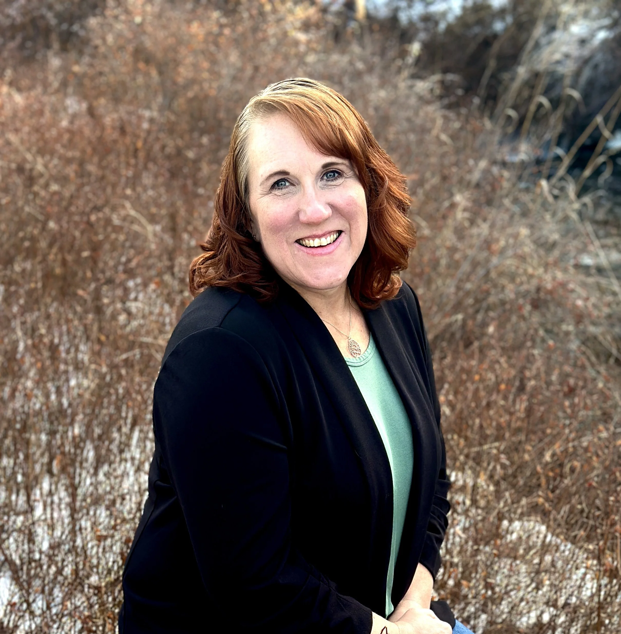 A woman with red hair smiling outdoors, wearing a black blazer over a light green top, with a background of autumnal foliage.