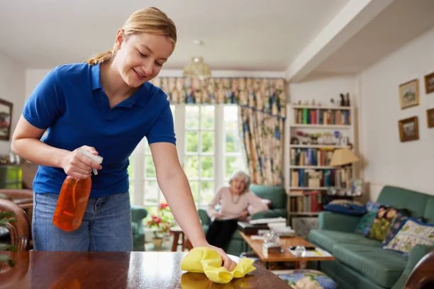 A woman cleaning a wooden table with a spray bottle and yellow cloth while a senior woman reads on a couch in a living room.