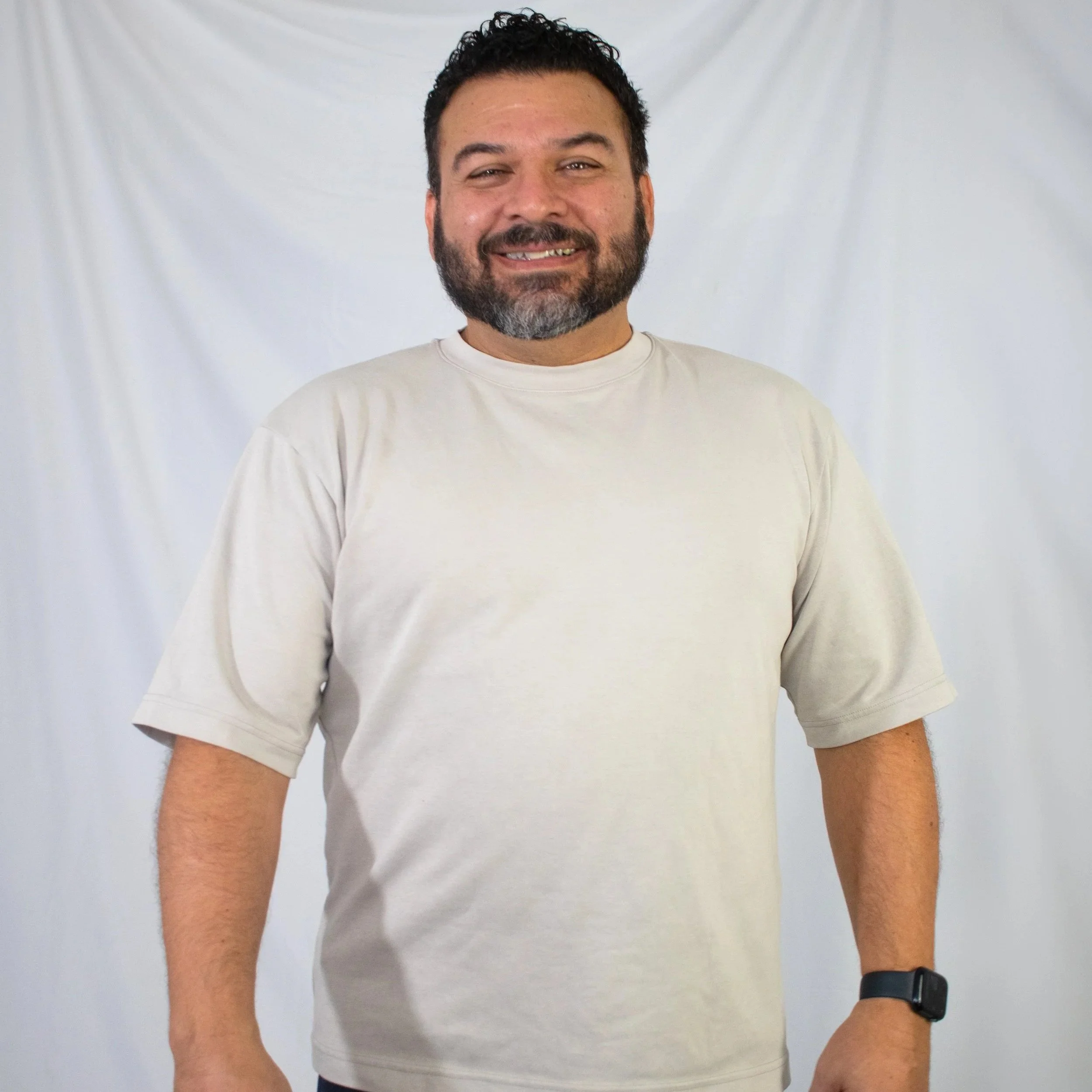 Close-up of a smiling man with dark, curly hair and a beard, wearing a white t-shirt and a black wristwatch, against a plain white background.