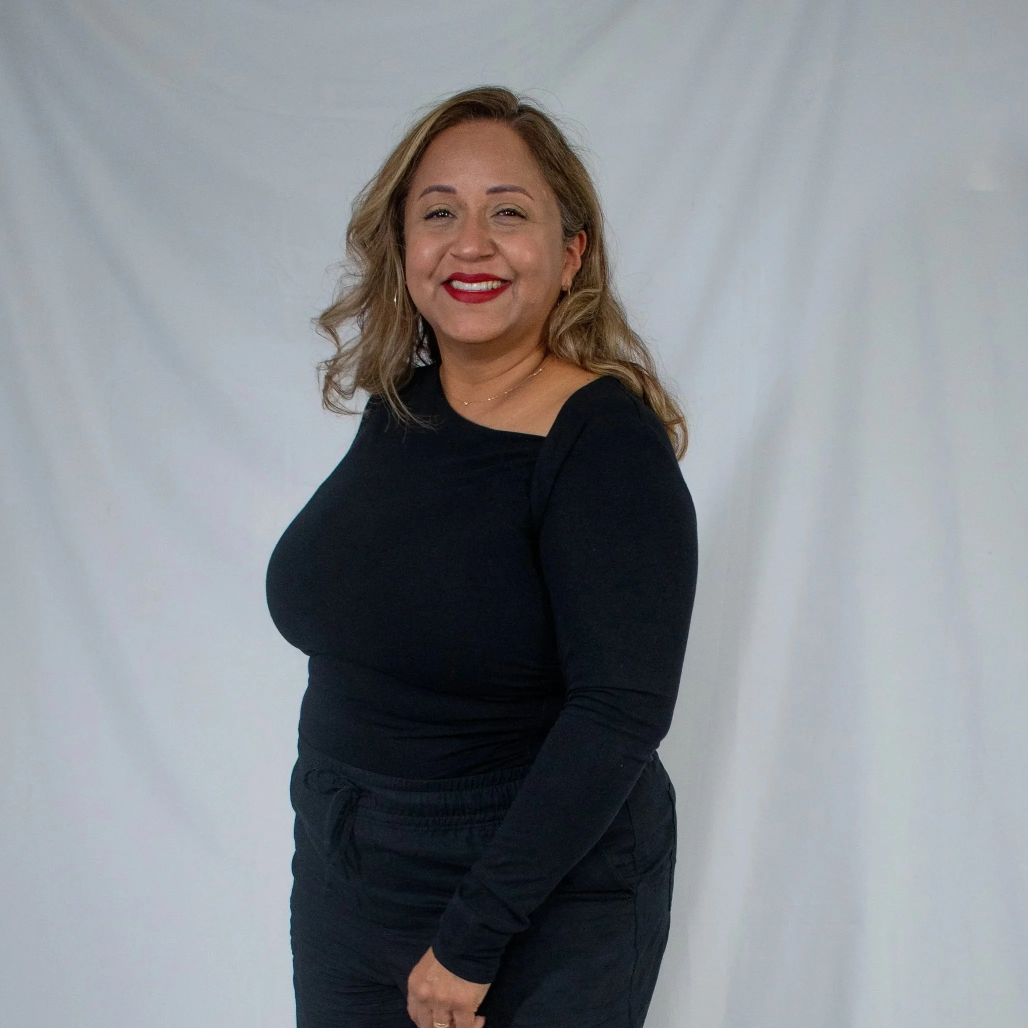A woman with wavy blonde hair, red lipstick, and hoop earrings smiling while posing against a plain light gray background. She is wearing a black top.
