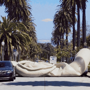 Large white reclining sculpture of a person lying down on a street with palm trees and a Hollywood sign in the background.