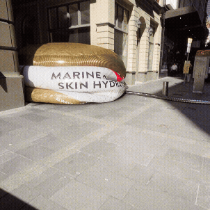 A large inflatable float resembling a skincare product labeled 'Marine Skin Hydra' is tied to a street pole outside a building.