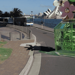 A tram or train on a track near a waterway, with a person walking along the sidewalk, a large green perfume display, and the Sydney Opera House in the background.