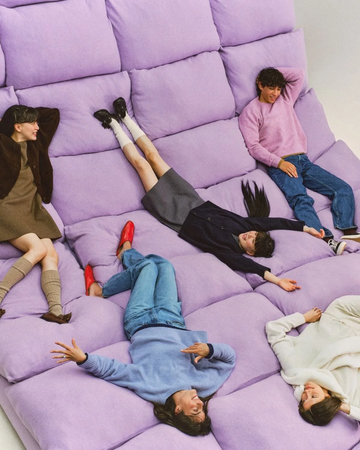 Five women in colorful sweaters and jeans relaxing and lying on a large purple sofa.