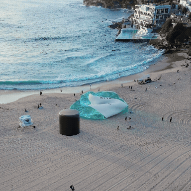 A large, round, beige structure with a lid on a sandy beach near the ocean, with a coastal town in the background.