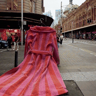 A pink and red striped coat draped over a stand on a city sidewalk with pedestrians and urban buildings in the background.