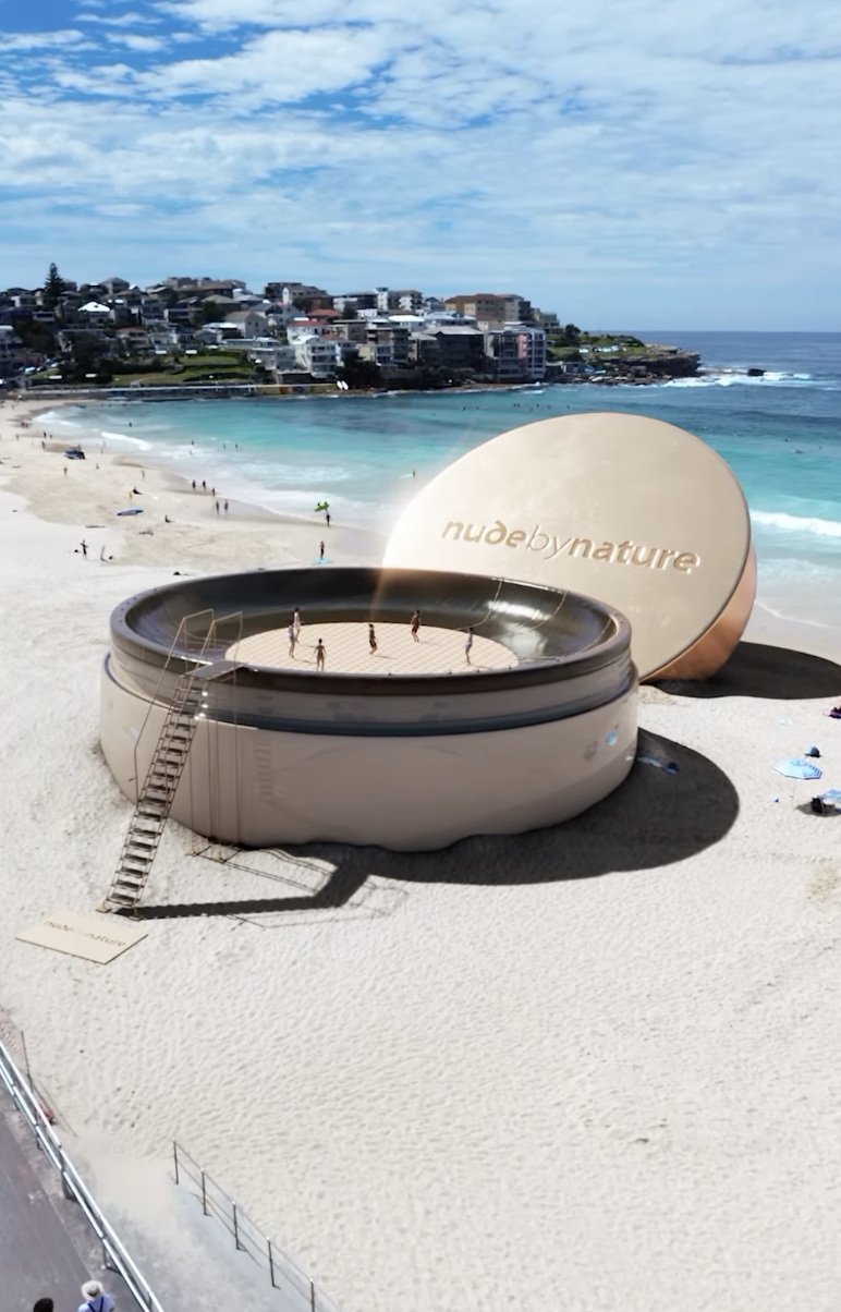 A large, round, inflatable hot tub on a sandy beach with a cover labeled 'nude by nature' leaning against it, and several people inside the hot tub, overlooking the ocean and a coastal town in the background.
