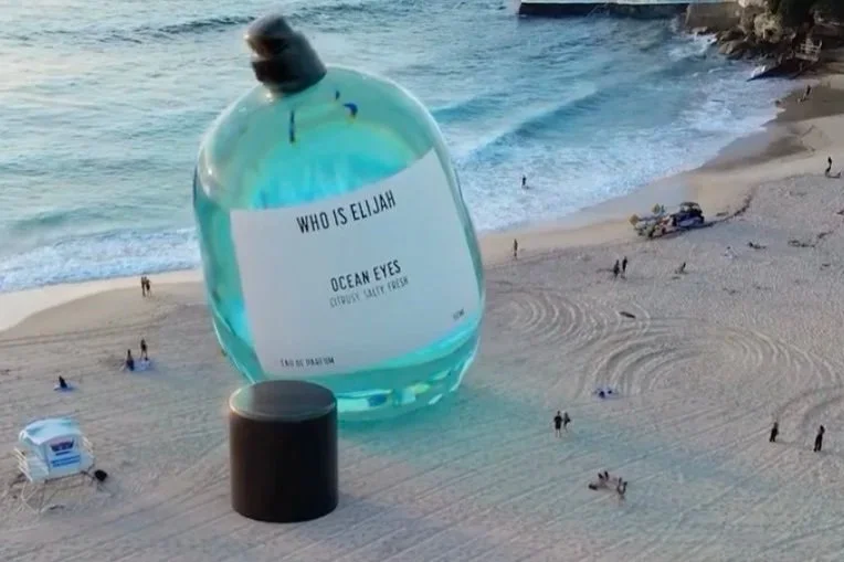 A giant perfume bottle labeled 'Who is Elijah' placed on a beach with people, beach chairs, and a shoreline in the background.