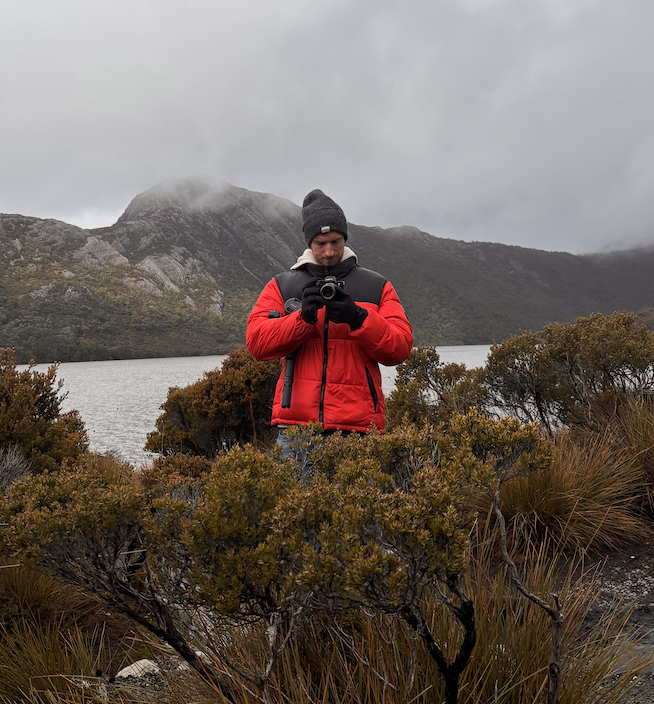 Person taking a selfie outdoors by a lake with mountains in the background, dressed in a red and black jacket and gray beanie, under cloudy skies.