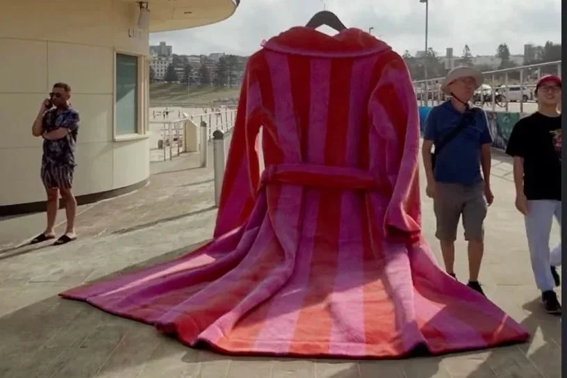 A large beach towel draped over a chair, making it look like a person is sitting in a towel outfit, with people walking by in an outdoor setting near a beach.