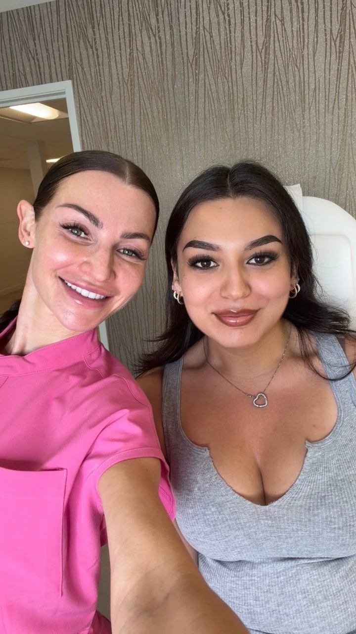 Two women taking a selfie indoors, smiling at the camera. The woman on the left is wearing a pink uniform, and the woman on the right is wearing a gray tank top with a jewelry necklace.