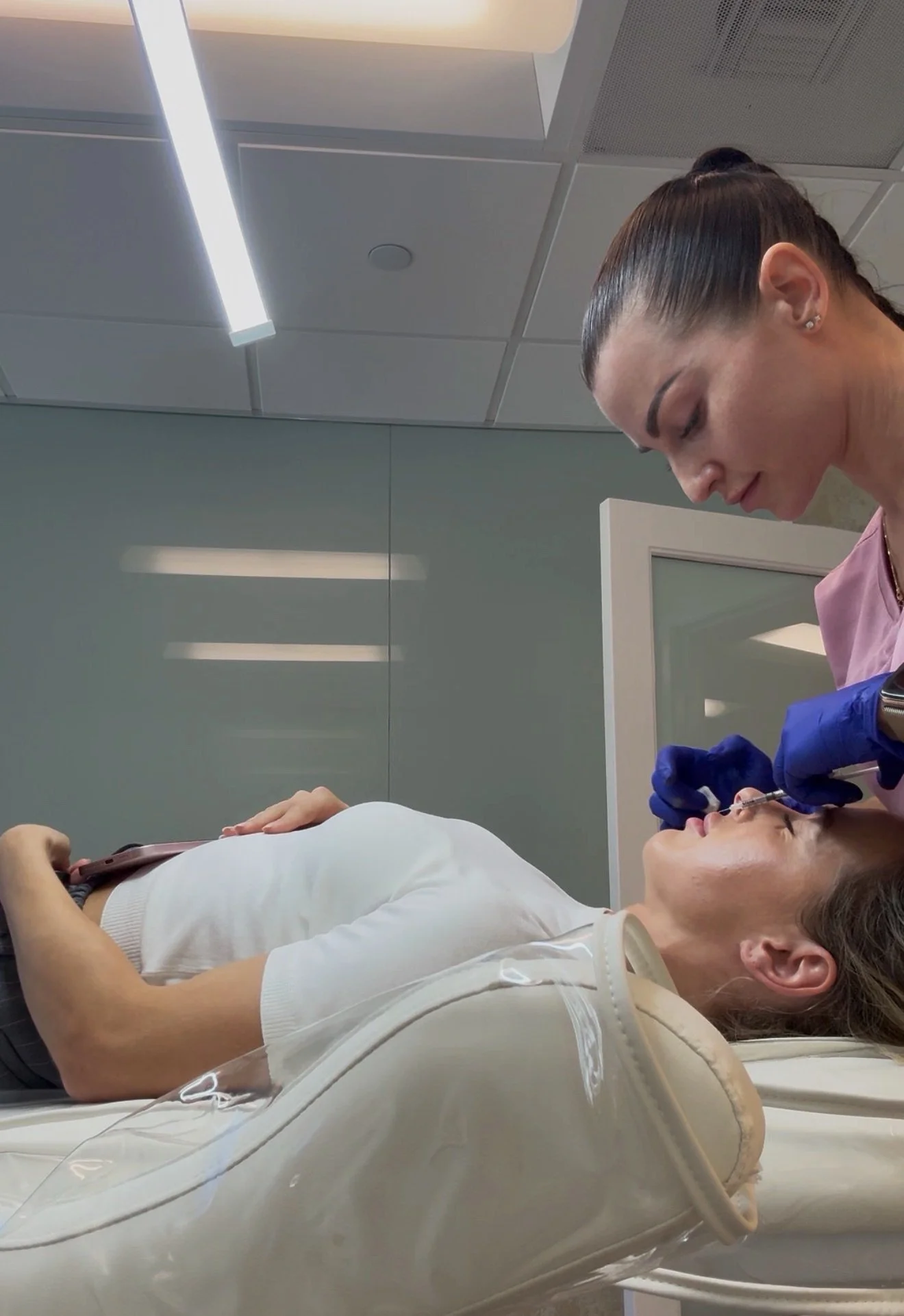 A woman lying on a medical chair receiving a cosmetic injection in her upper lip from a healthcare professional wearing blue gloves, in a clinical setting.