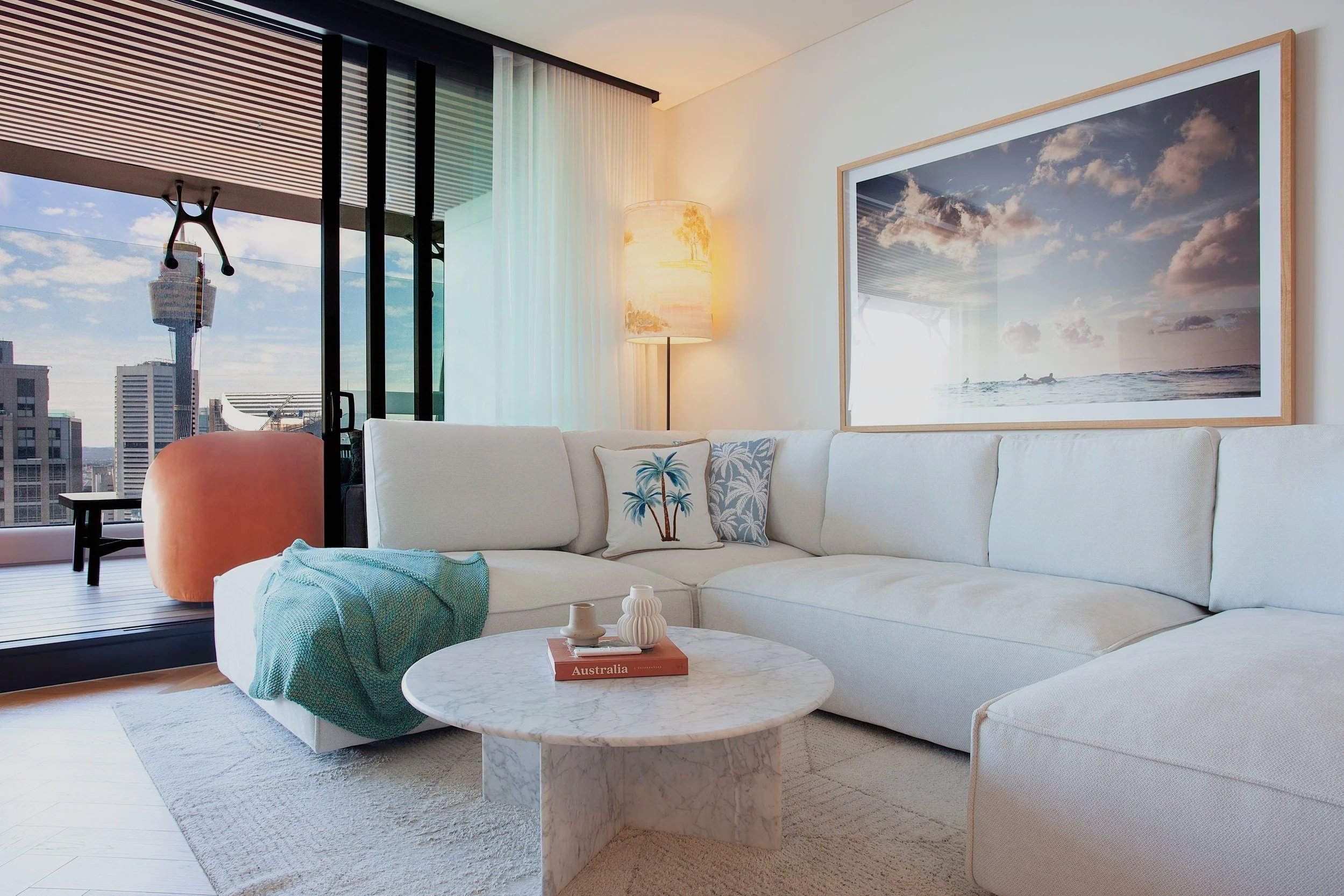 Modern living room with white sectional sofa, decorative pillows, marble coffee table, and wall art of a beach scene. Large window with city skyline view, curtains, and a floor lamp with a lampshade near the corner.