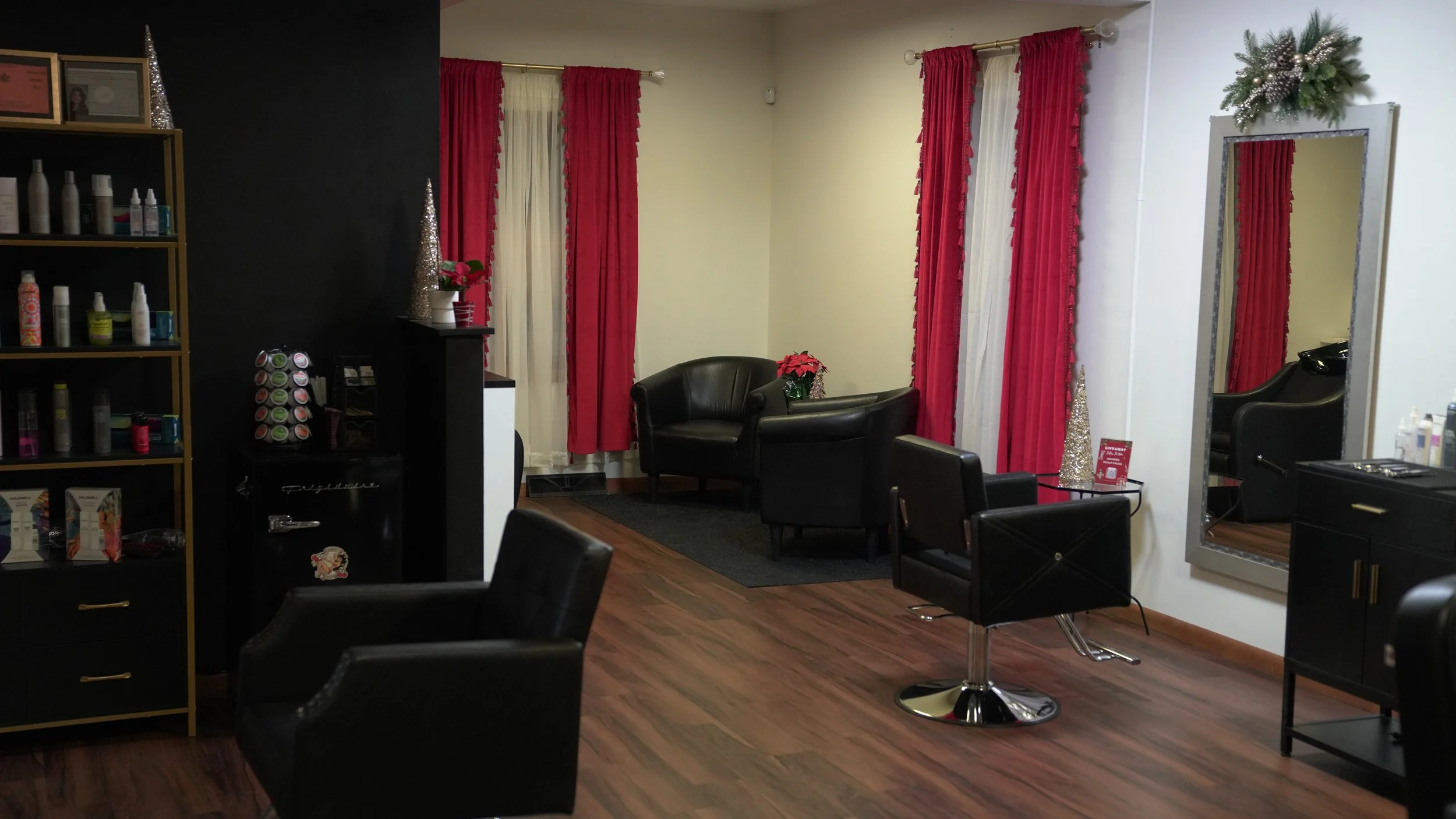 Interior of a beauty salon or hair salon, featuring black chairs in stylist stations, a mirror, and a shelf with hair products, with festive Christmas decorations including poinsettias, pinecones, silver trees, red and white curtains, and a small black table with a sign.
