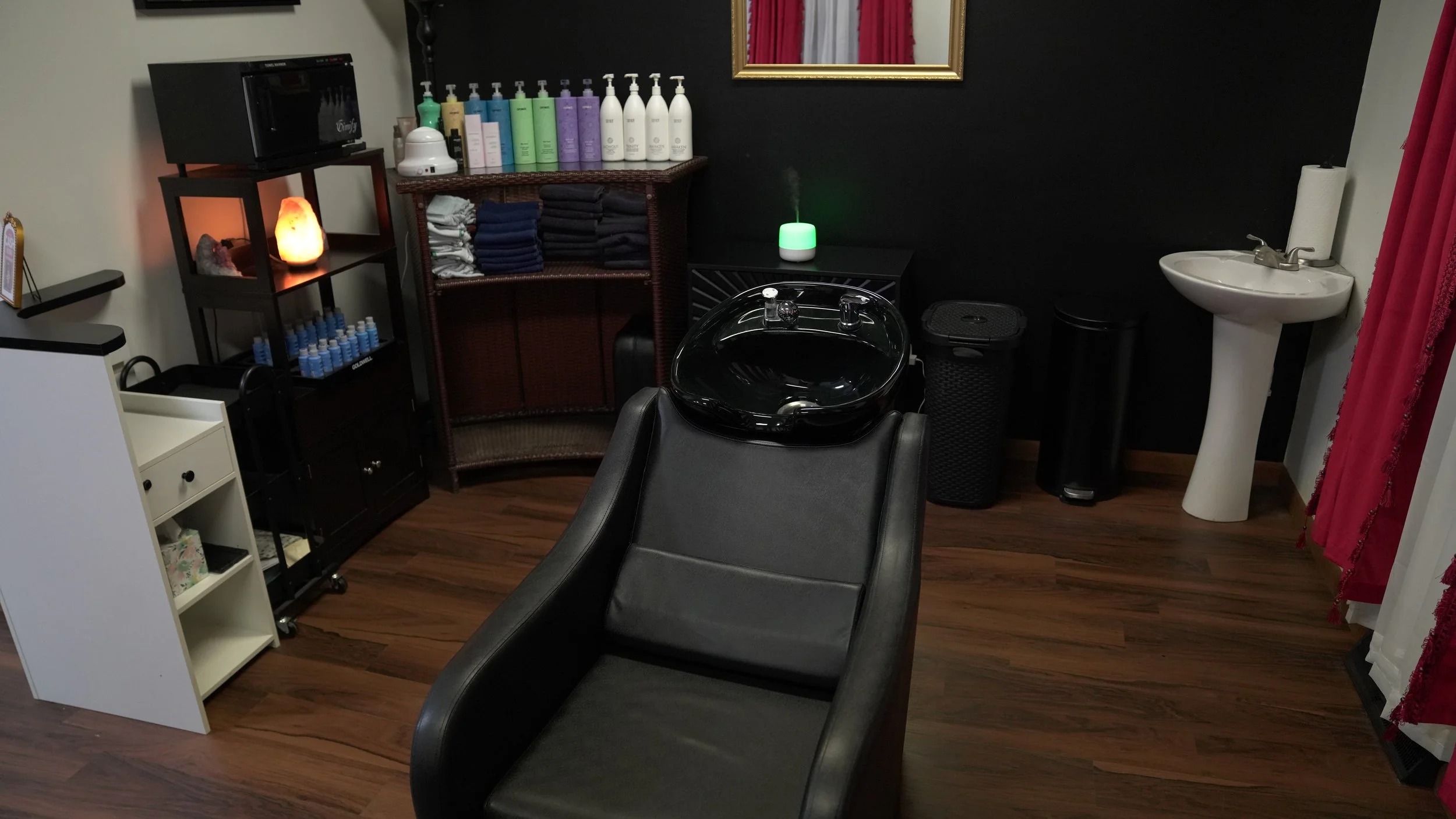 A salon or spa treatment room with a black shampoo basin on a reclining chair, a shelf with towels and hair products, a small sink with soap and paper towel dispenser, a diffuser emitting vapor, a salt lamp, and nail polish bottles, with a framed mirror on a black wall and red curtains on the side.