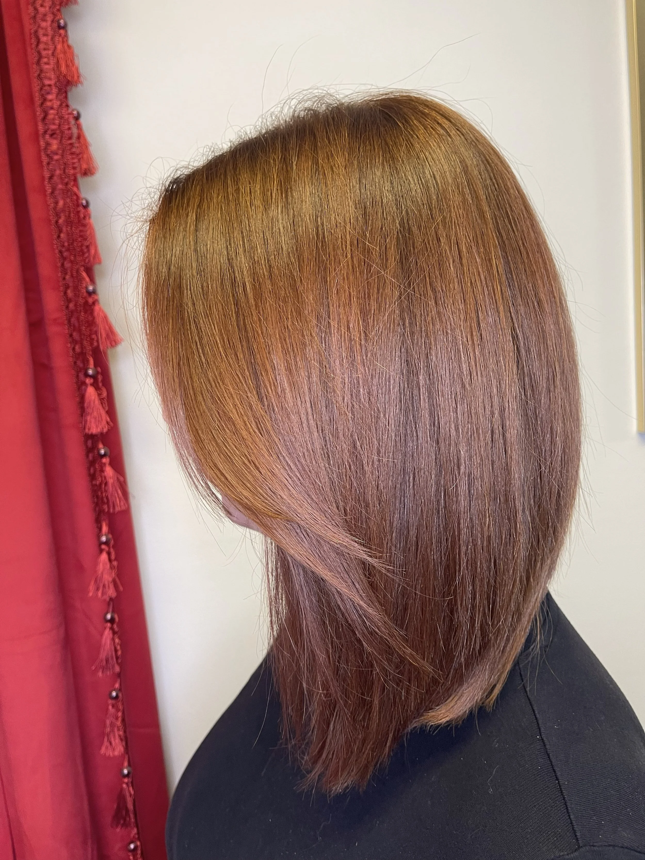 Side view of a woman's head with straight, shoulder-length, shiny reddish-brown hair. She is wearing a black top and standing near a white wall with a red curtain on the left side.